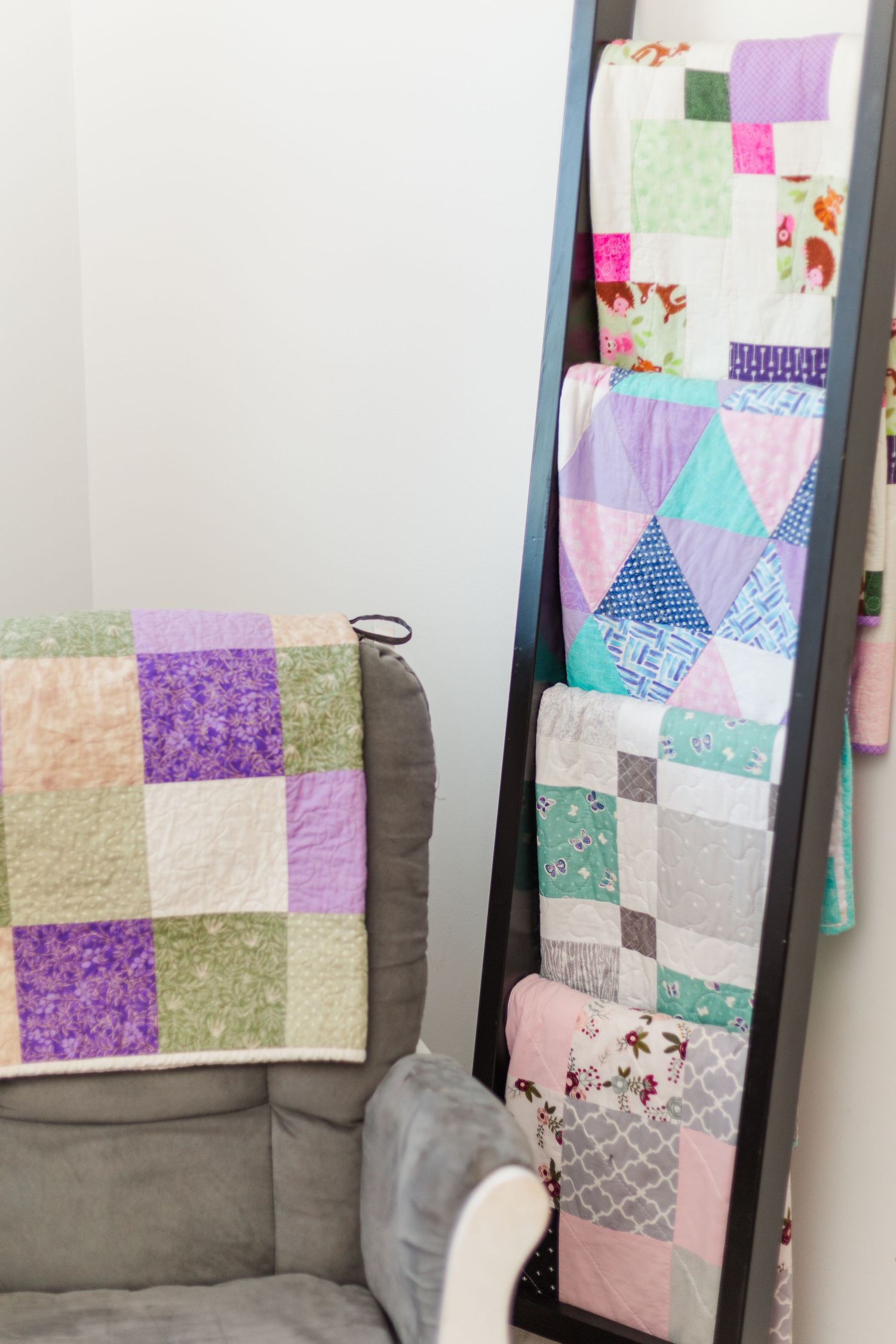 Handmade patchwork quilts displayed on a gray couch and leaning against a wall, featuring purple, pink and teal patterns.