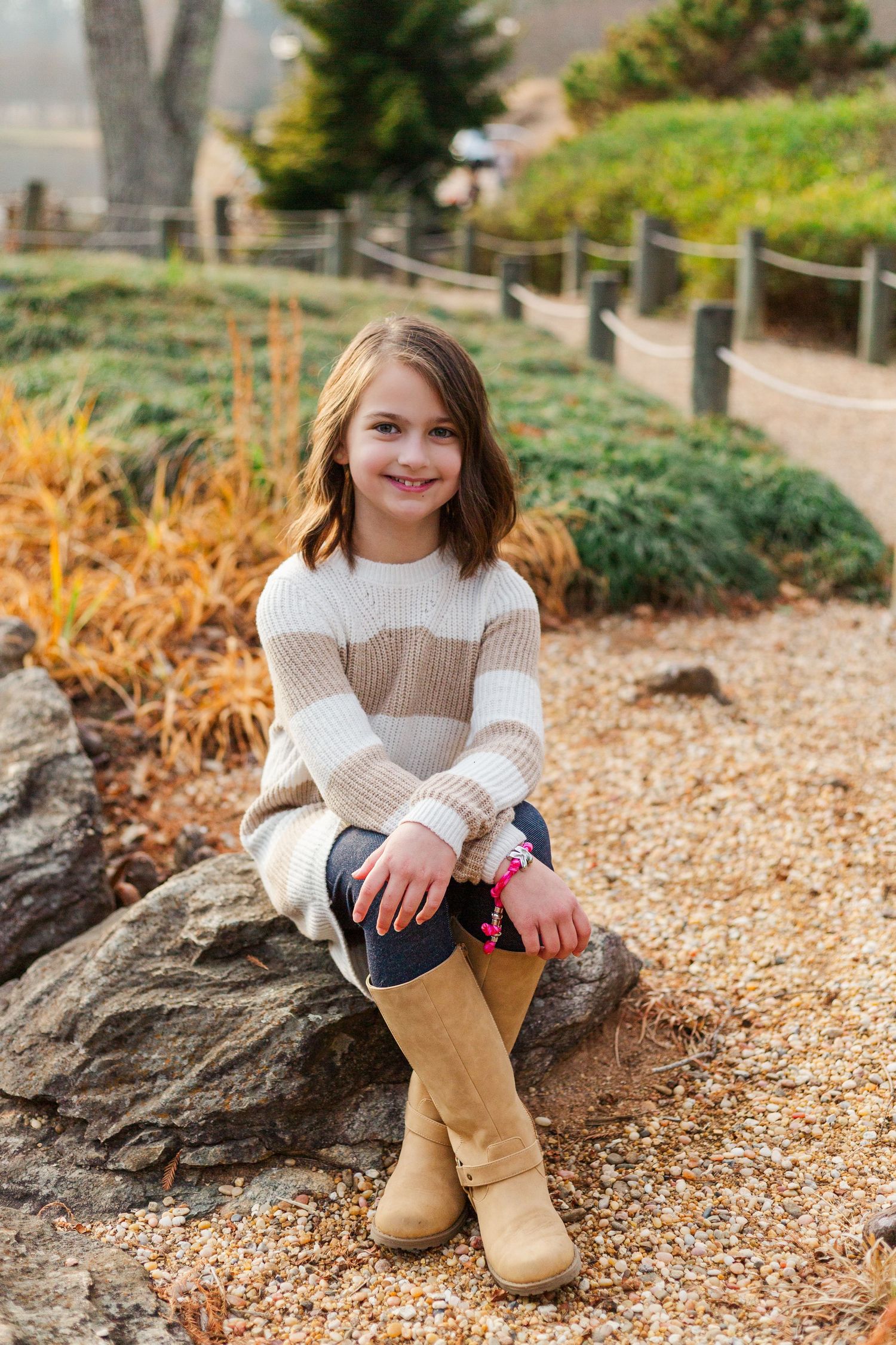 A child in a white knit sweater sits on a rock path surrounded by autumn foliage and landscaping.