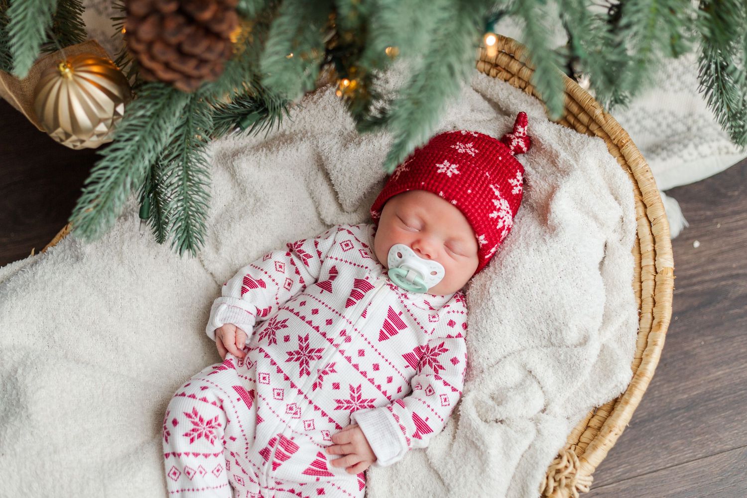 Baby wearing red winter hat and festive holiday pajamas sleeps in woven basket with pinecone Christmas decorations.