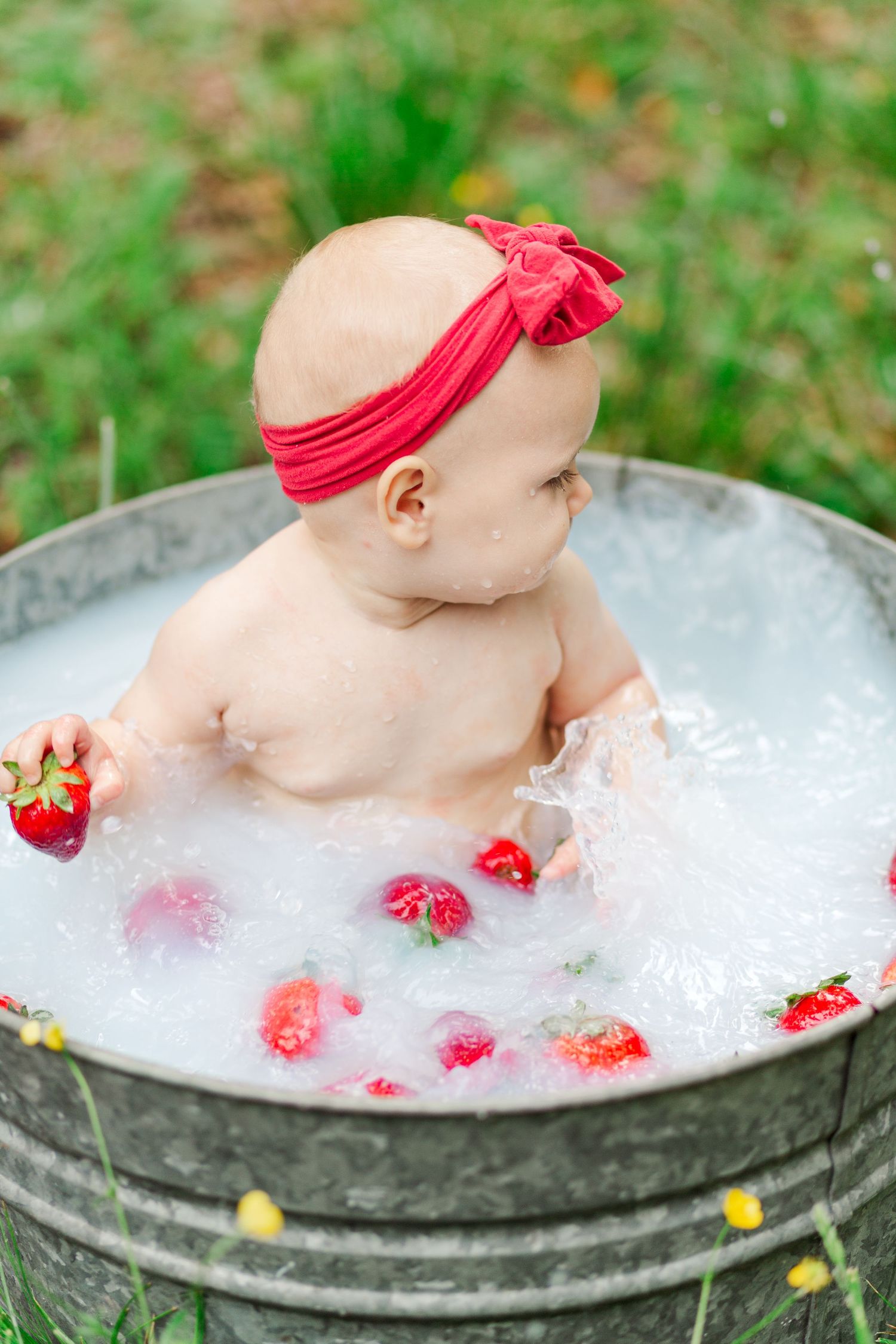 Baby in a metal tub filled with bubbles and strawberries wearing a red headband with bow for summer photoshoot in Greenville, SC