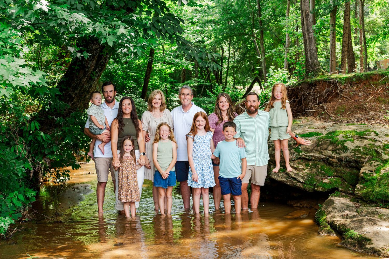 A group plays and splashes in a shallow creek surrounded by lush green forest on a summer day.