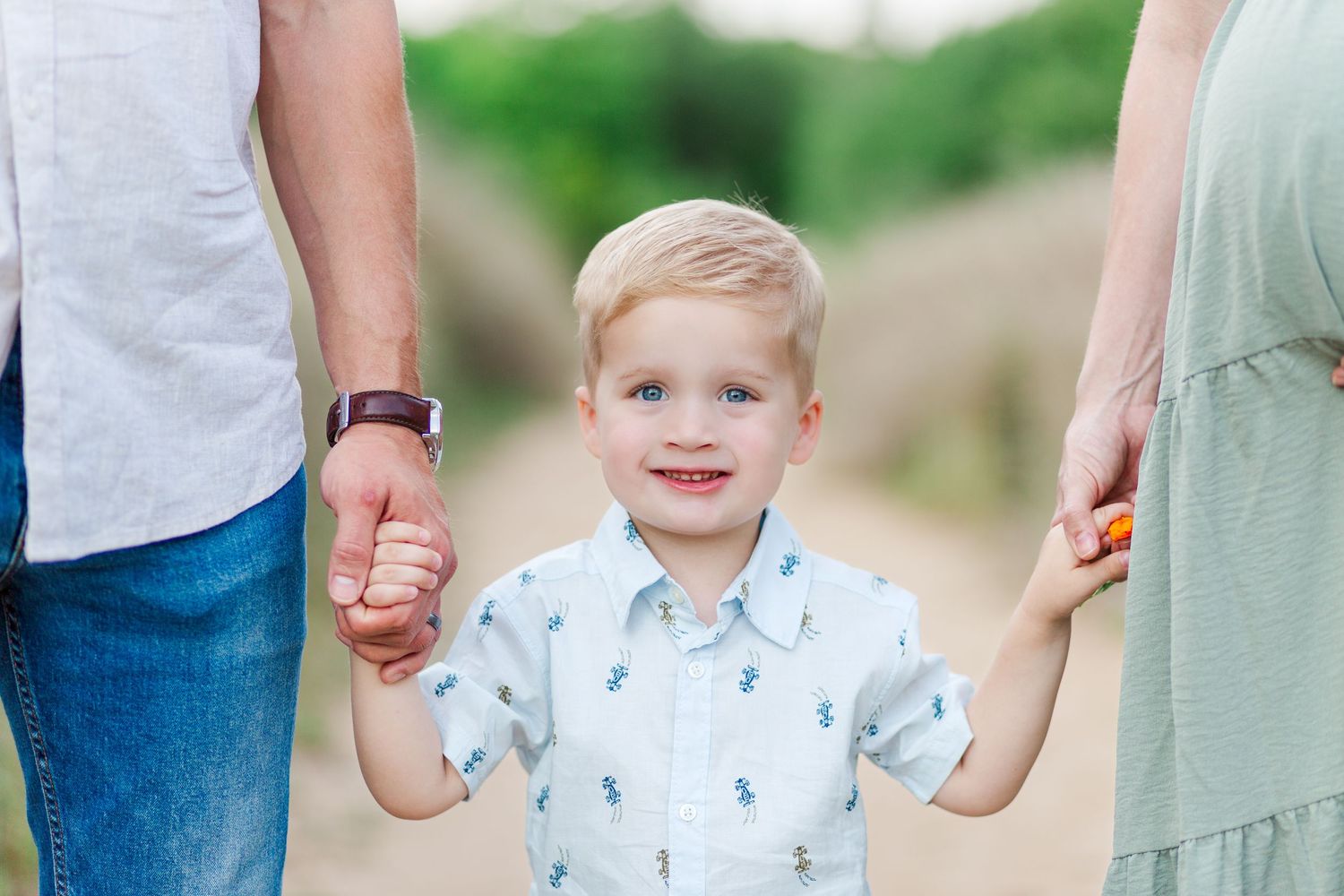 Toddler wearing white outfit in Clemson, SC holds hands with parents as they walk through a grassy outdoor setting for family photo session.
