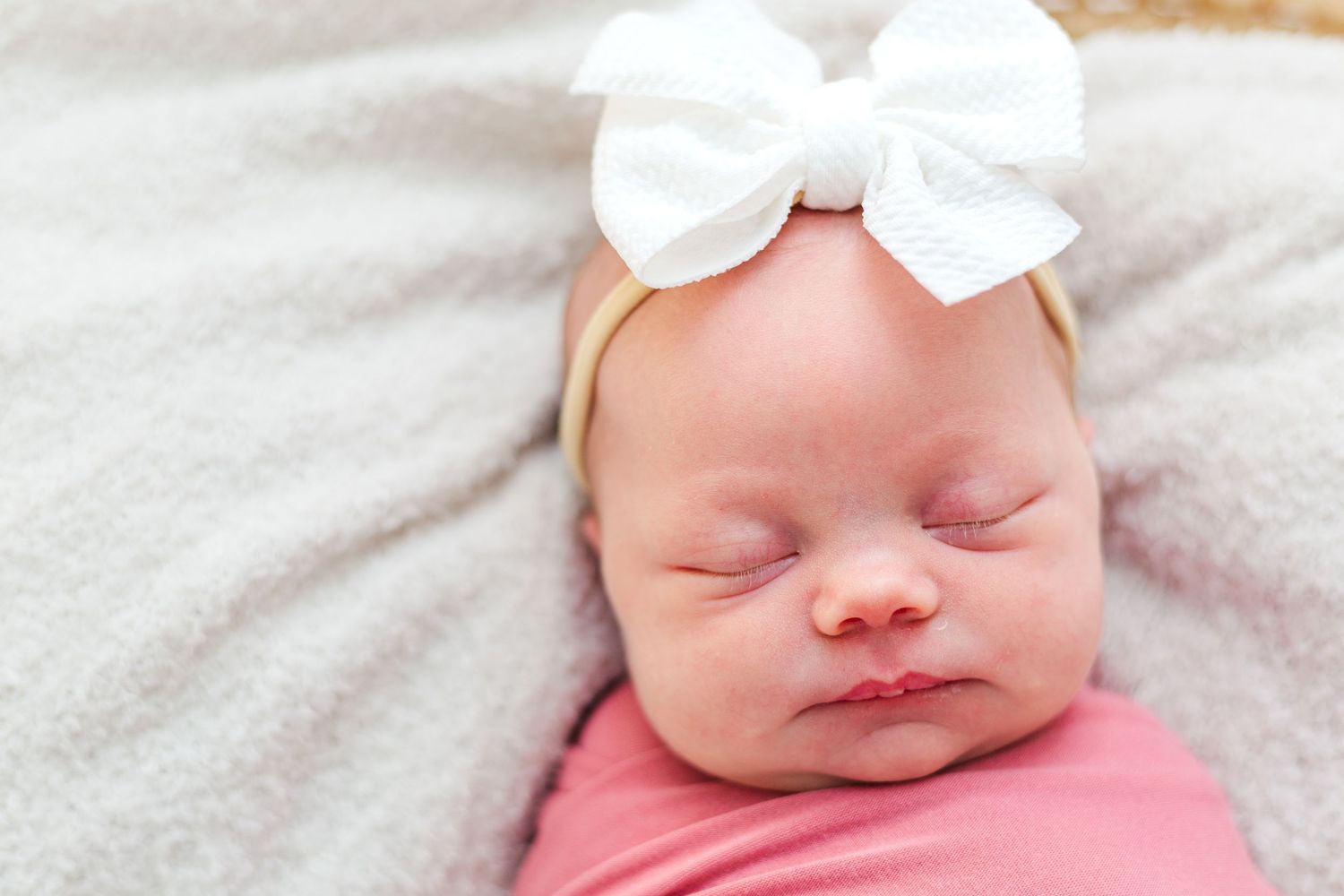 Peaceful newborn baby sleeping peacefully on white blanket wearing pink clothing and white bow headband.