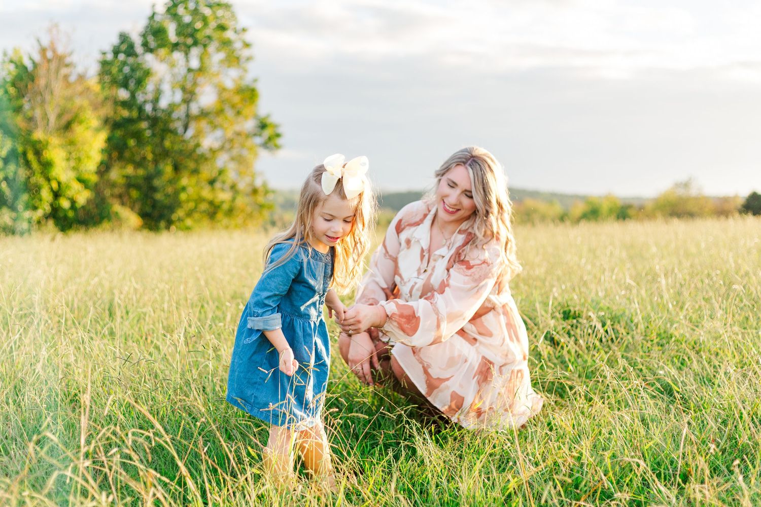 A mother and young daughter in a floral dress and denim outfit playing in a sunny green meadow.