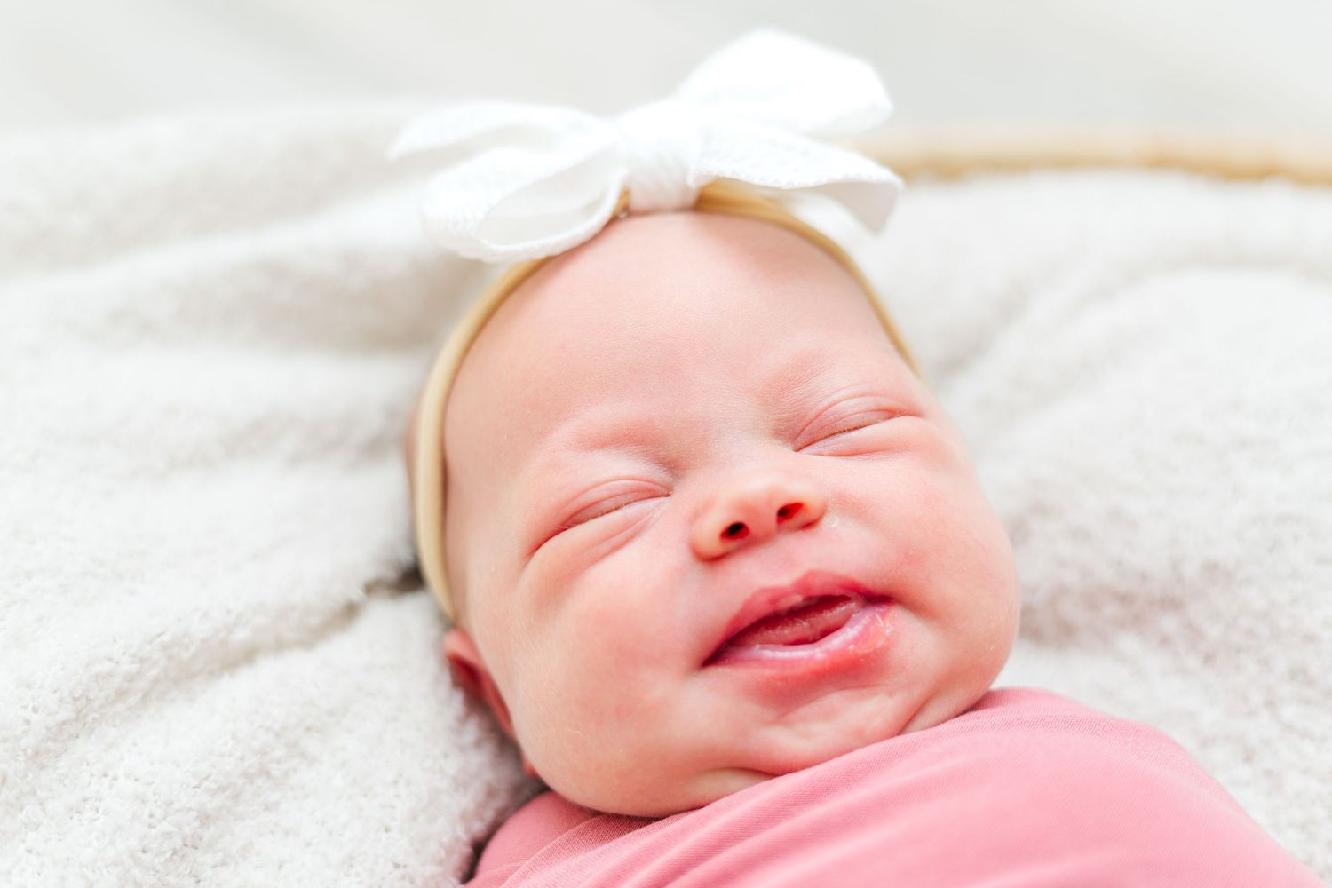 A newborn baby wearing a white headband smiles while lying on a soft blanket.