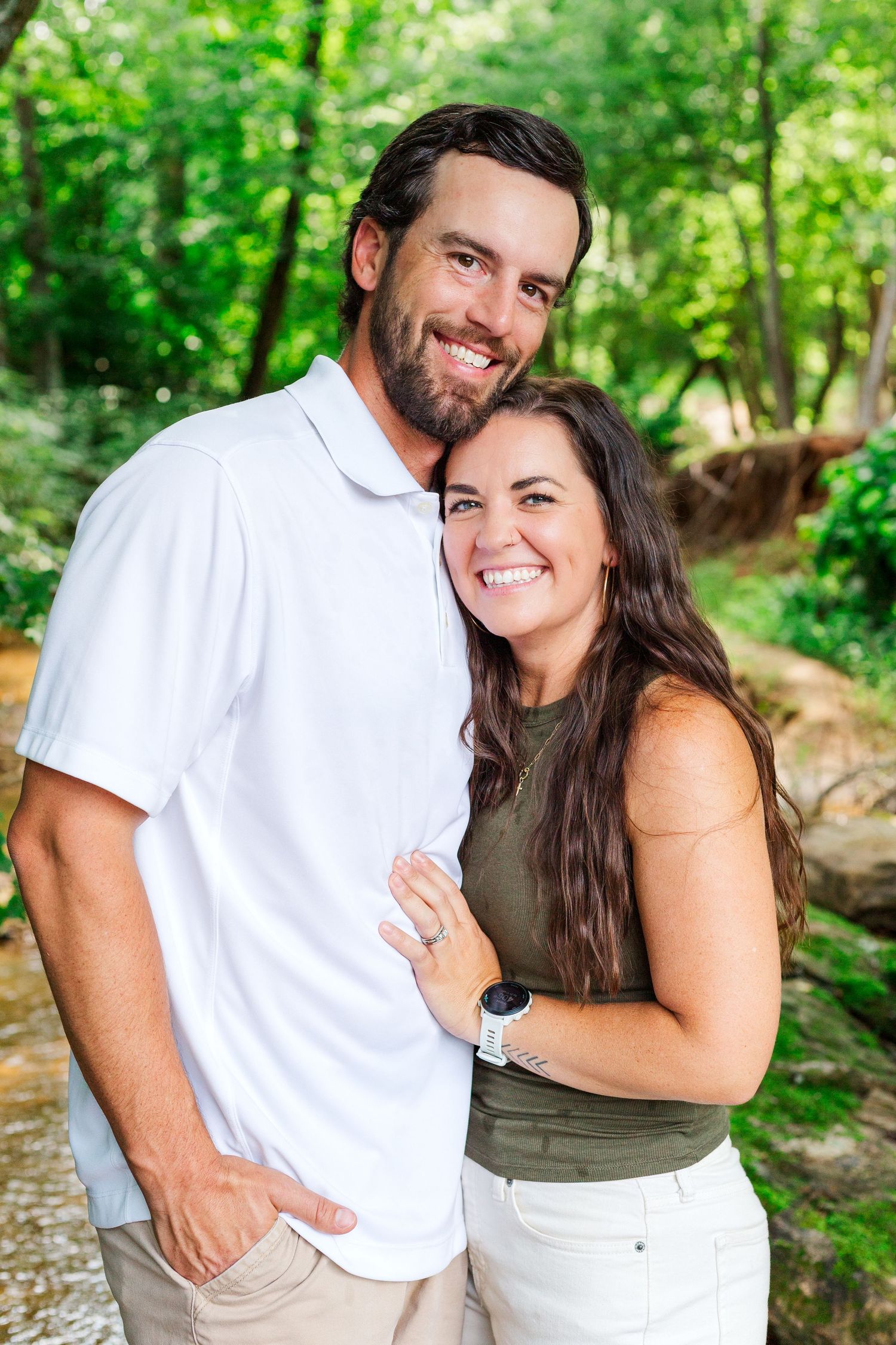 A couple poses together outdoors in a wooded setting with natural greenery in the background.