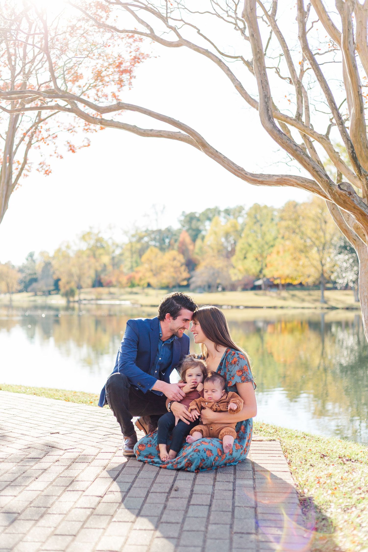 Family enjoying a tender moment together while sitting on a brick pathway by a serene autumn lake.