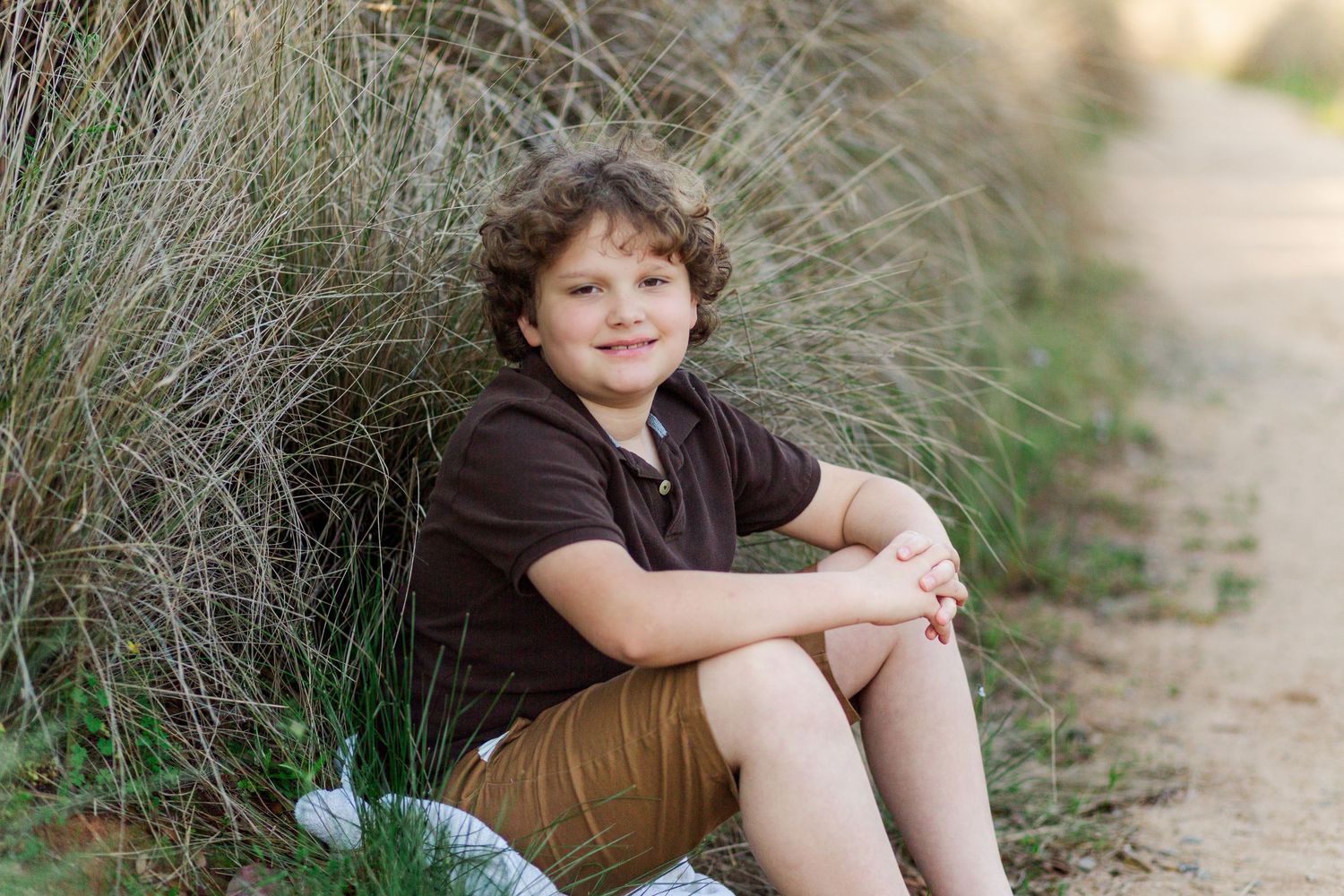 A young person in a brown shirt sits on the ground against tall grass on a sunny day.