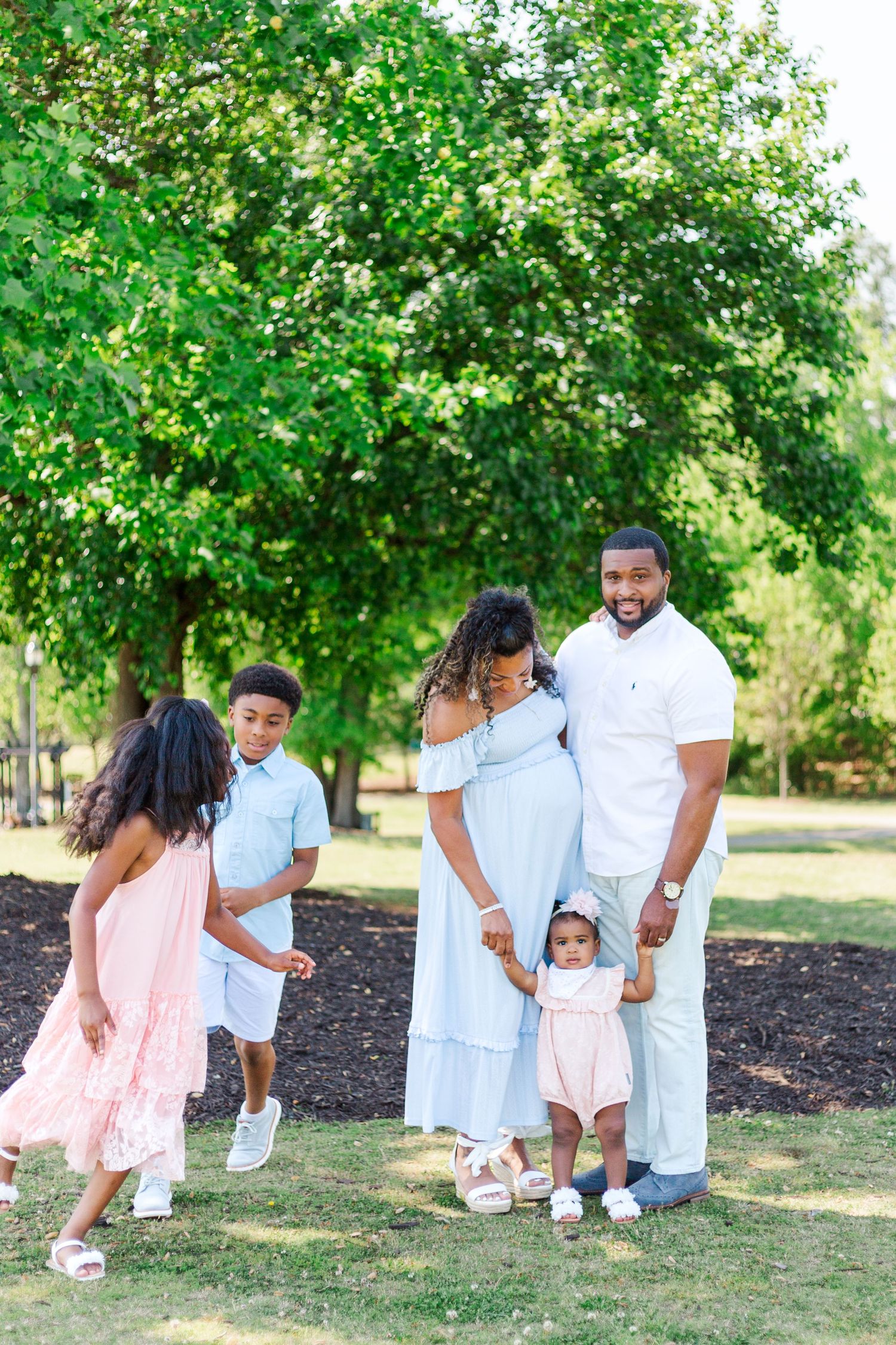 A joyful family poses outdoors in a lush green park, wearing coordinated pastel outfits, with a pregnant mother and toddler.