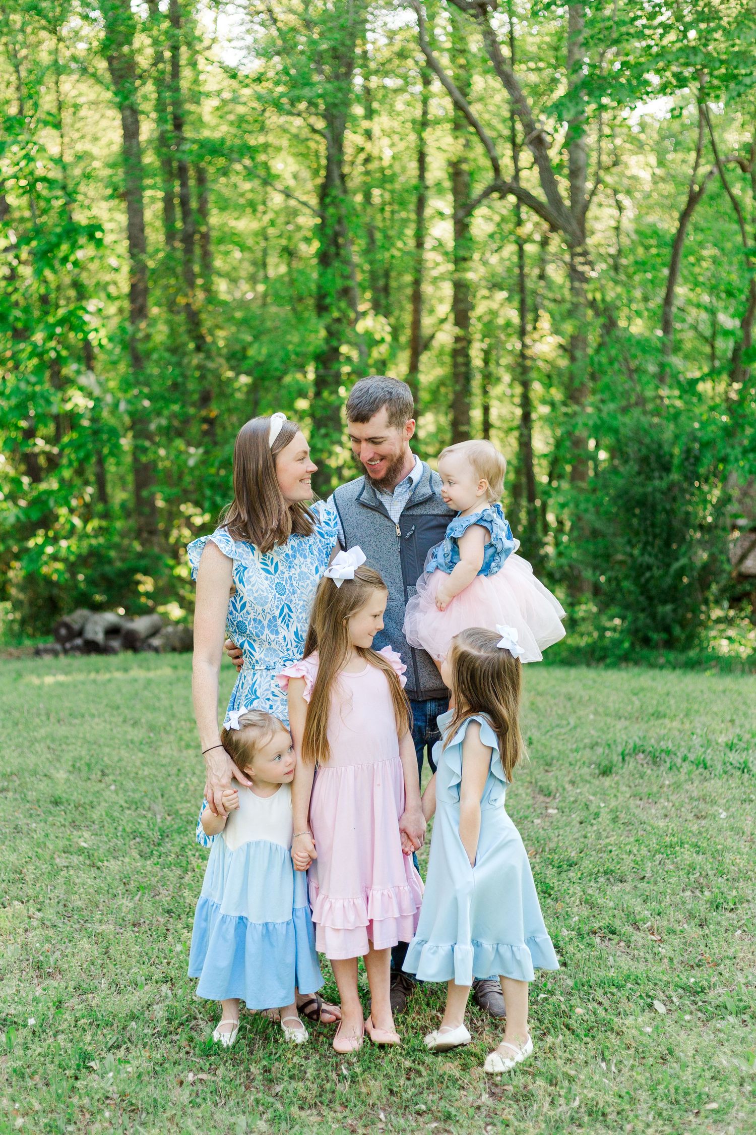 A happy family of six poses outdoors in coordinating pastel outfits, surrounded by lush green trees.