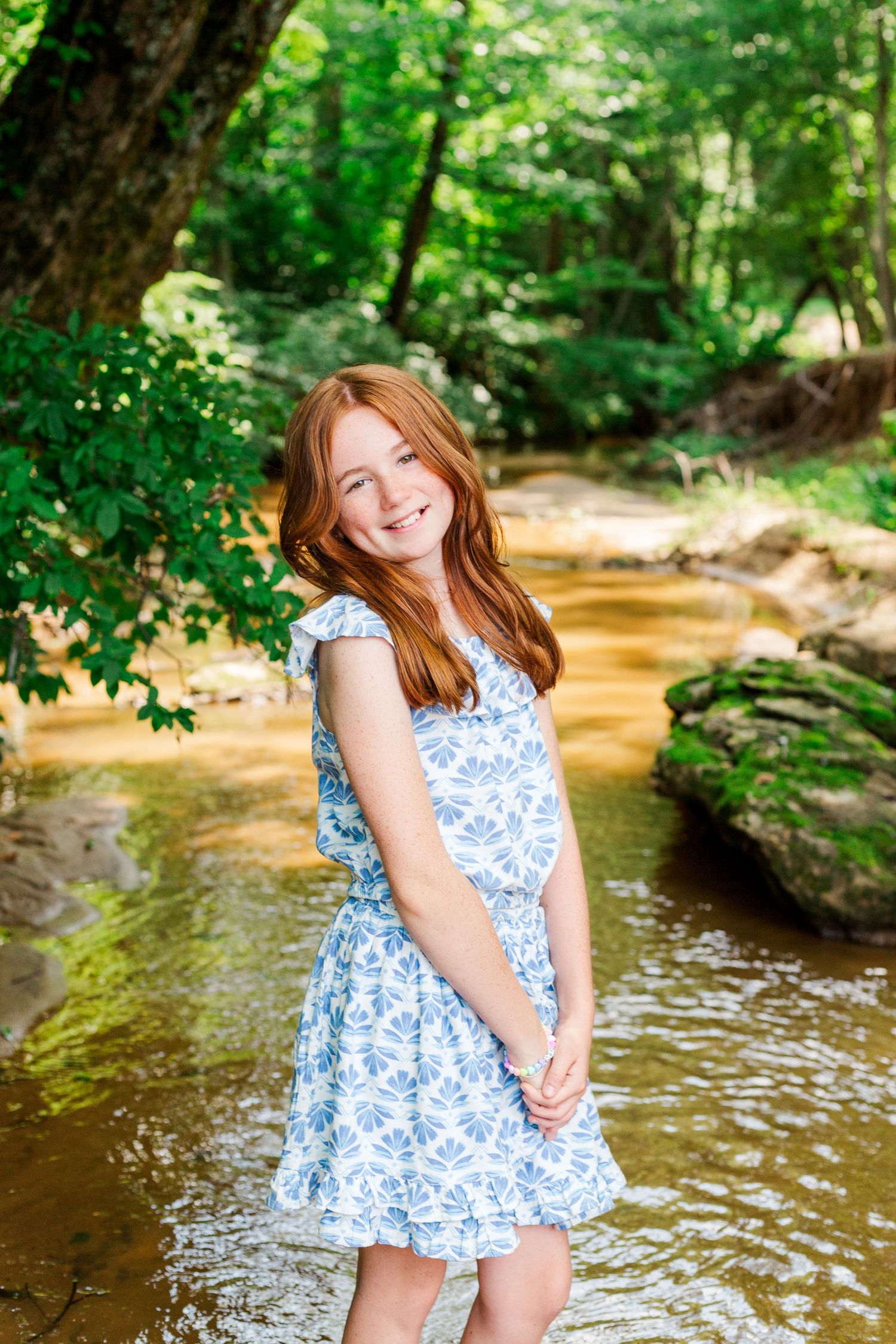 A young person in a blue floral dress poses in a sunlit creek with green forest scenery.
