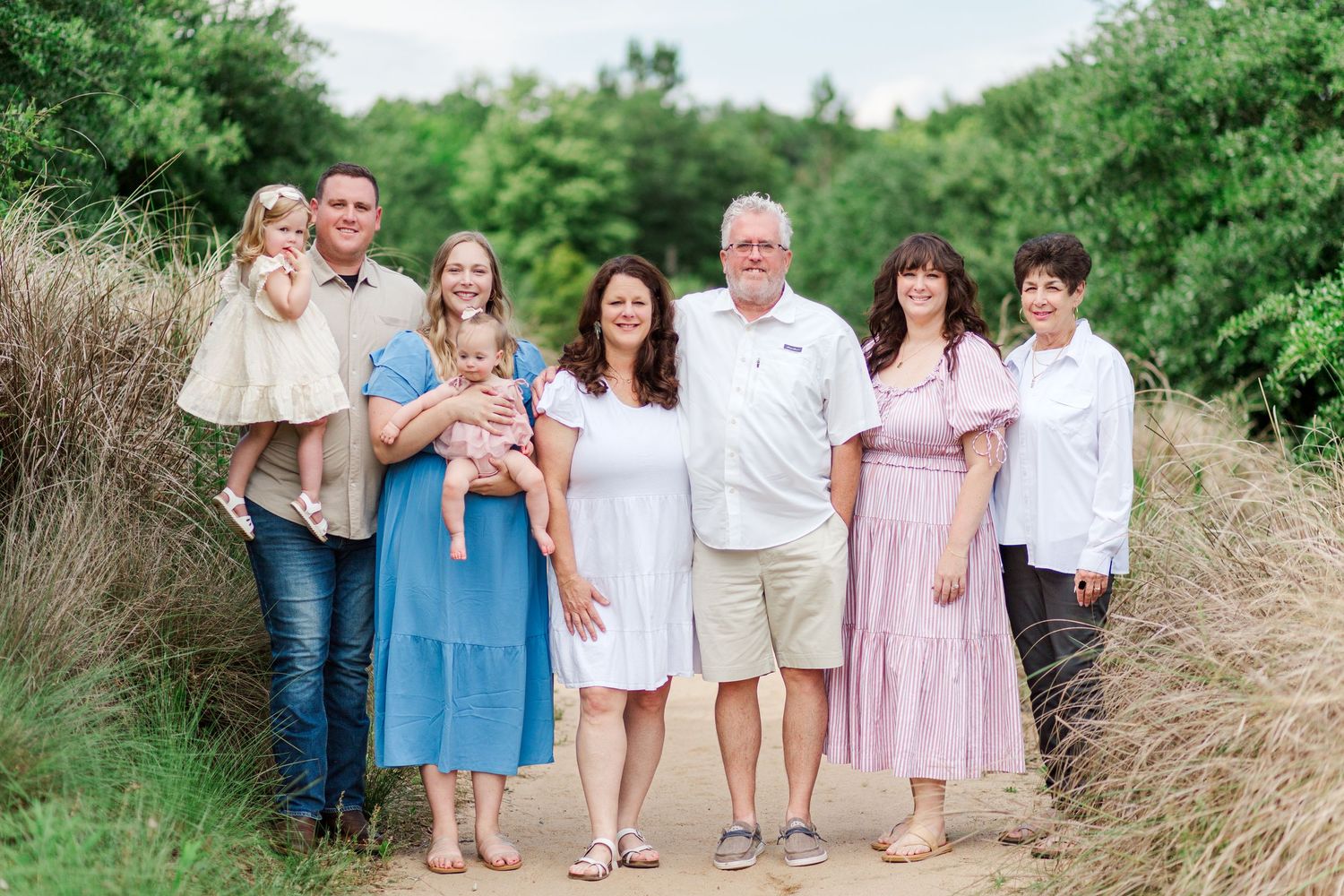 Three generations of a family in coordinating pastel outfits pose together on a grassy path during summer.