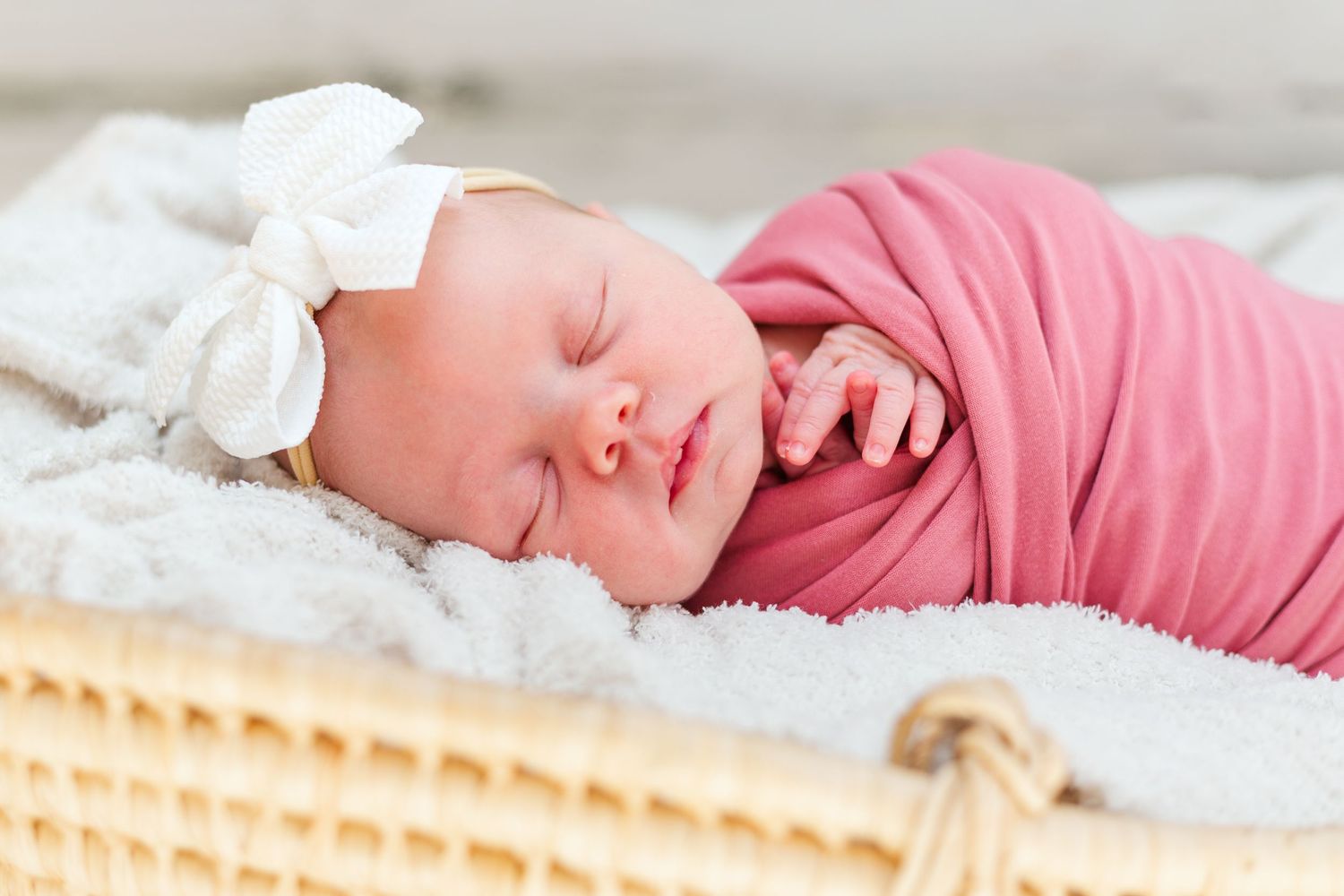 Newborn baby girl swaddled in pink wrap sleeping in a wicker basket with a white bow headband.
