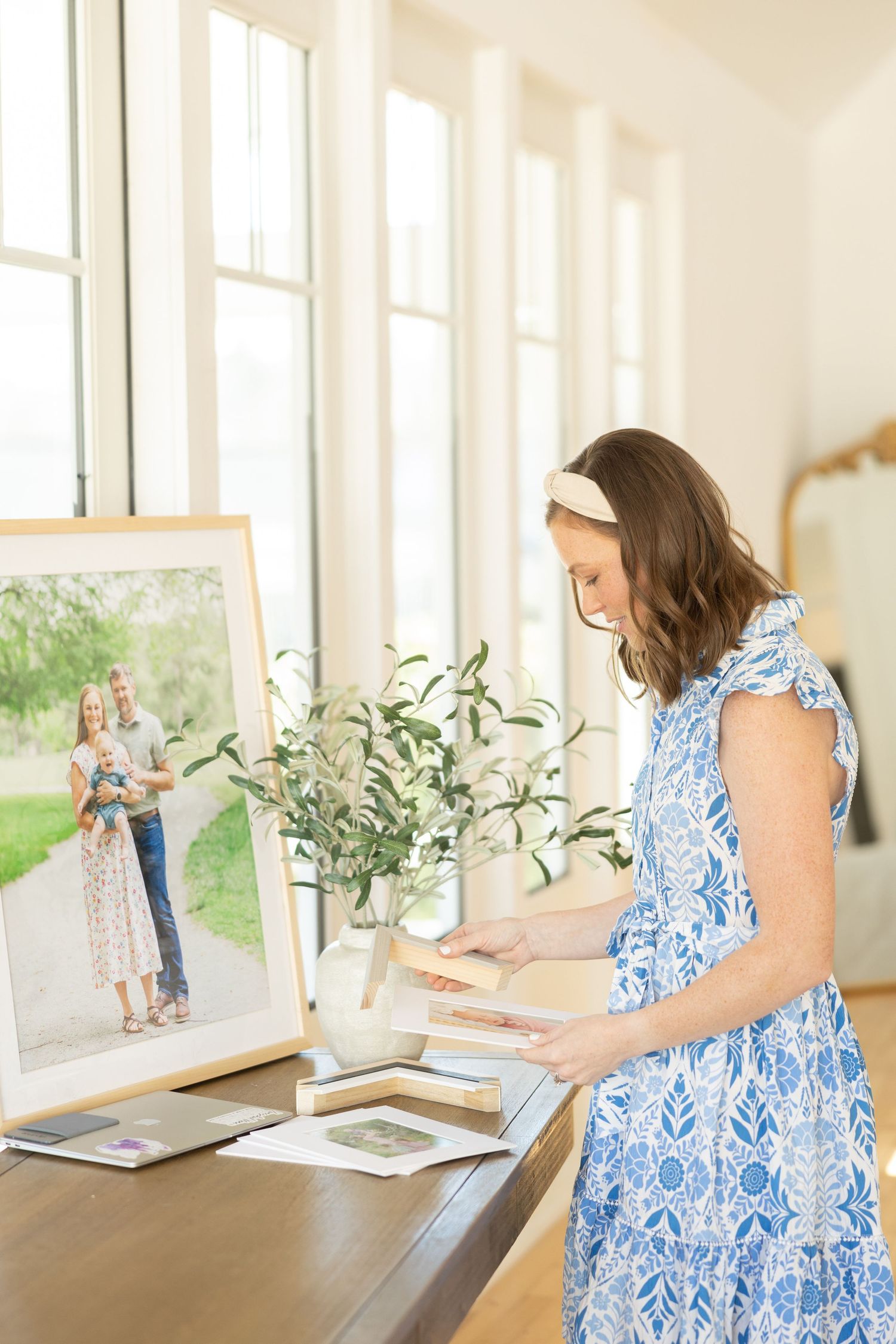 Woman in blue and white patterned dress reviews photographs displayed in bright sunlit room with plants.