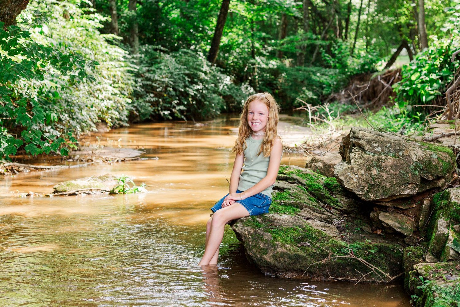A peaceful creek scene in the woods with mossy rocks and reddish-brown water surrounded by lush green foliage.