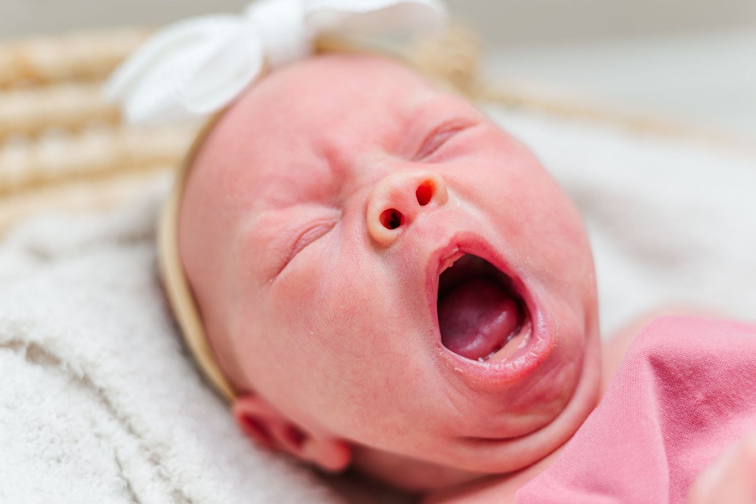 Close up of a yawning newborn baby wearing pink clothing while resting on a white textured surface.