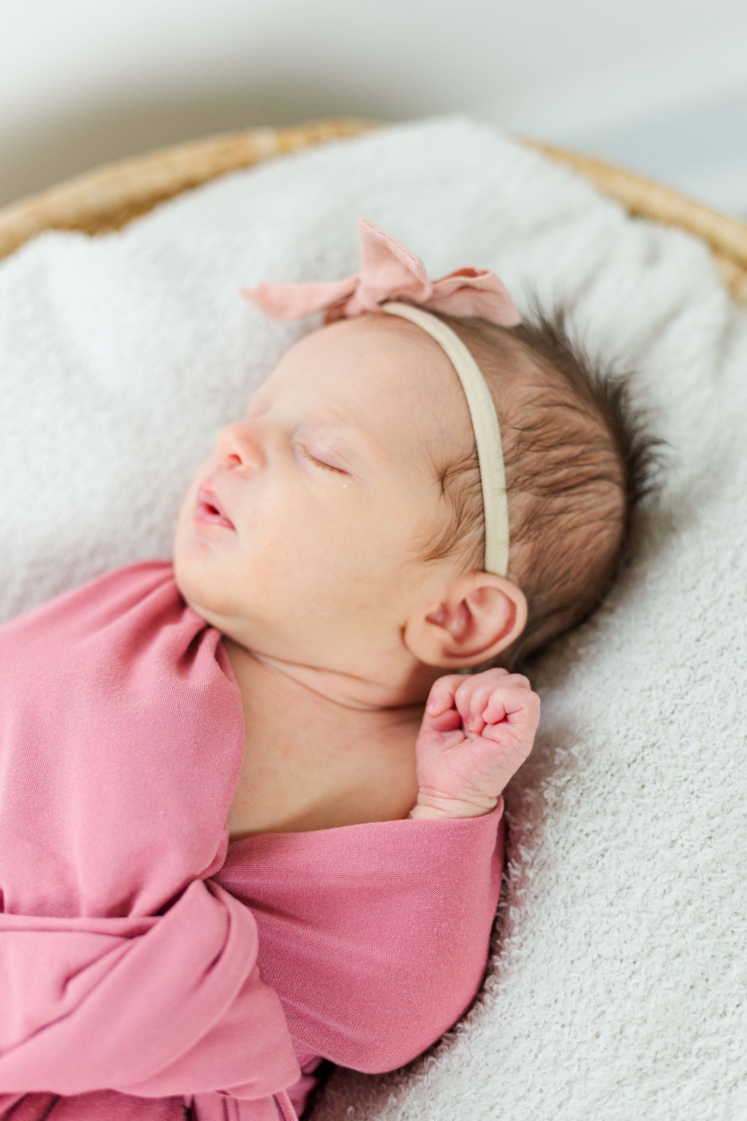 Sleeping newborn baby girl wrapped in pink swaddle with bow headband in wicker basket.