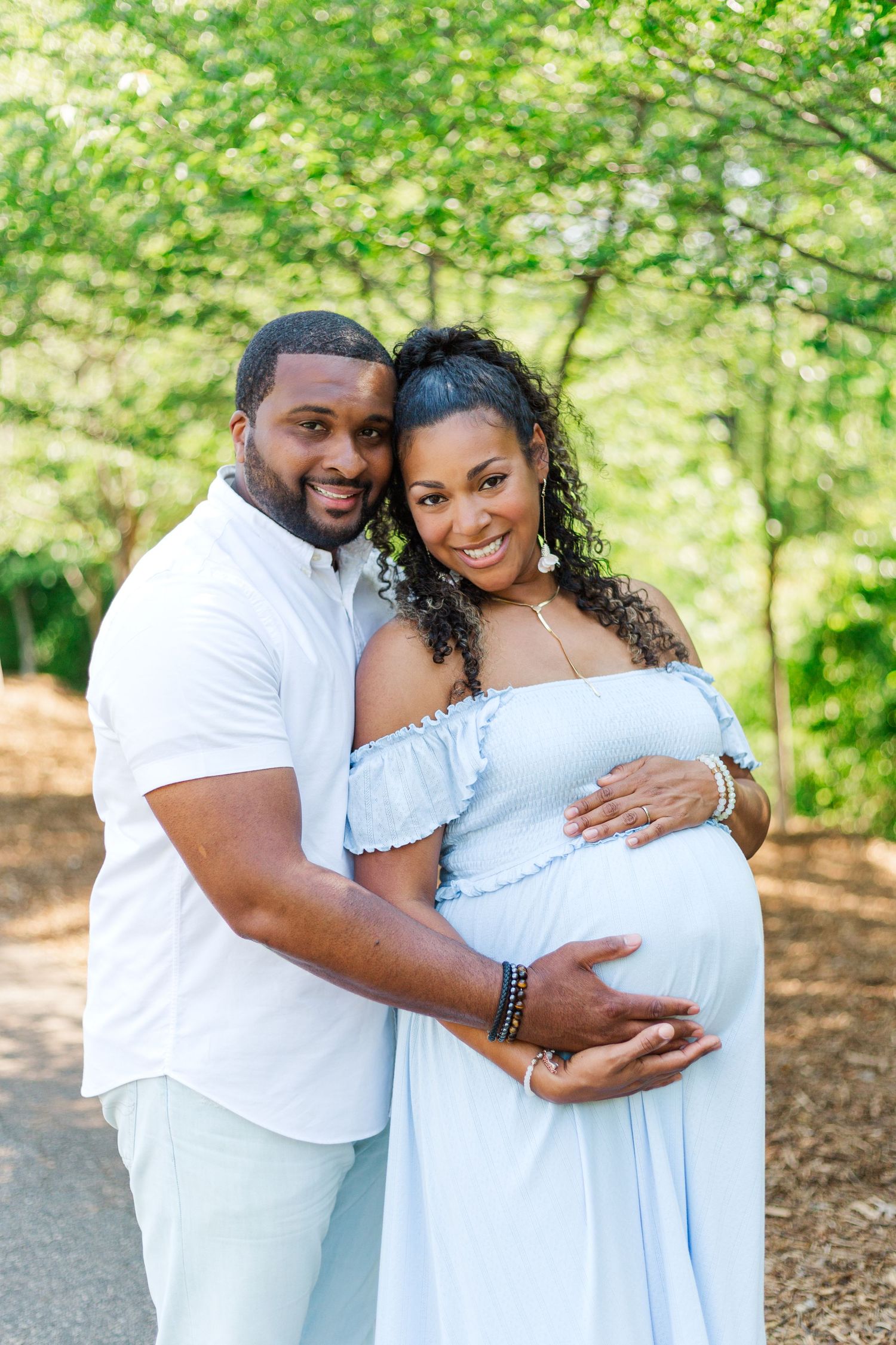 A smiling couple poses outdoors, the woman in a light blue maternity dress, both cradling her baby bump.