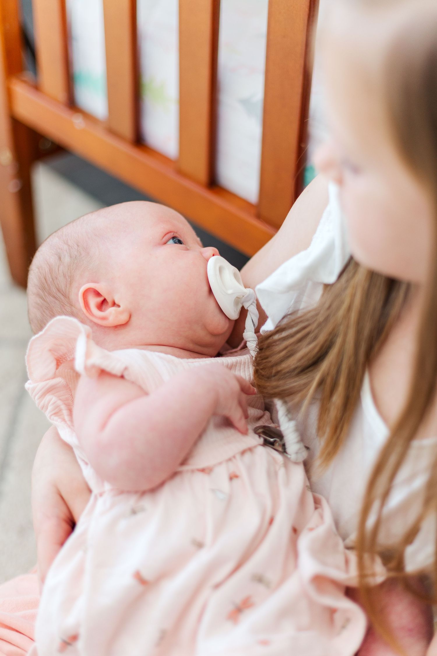 Baby wearing a soft pink dress looks up while being bottle fed near a wooden crib.