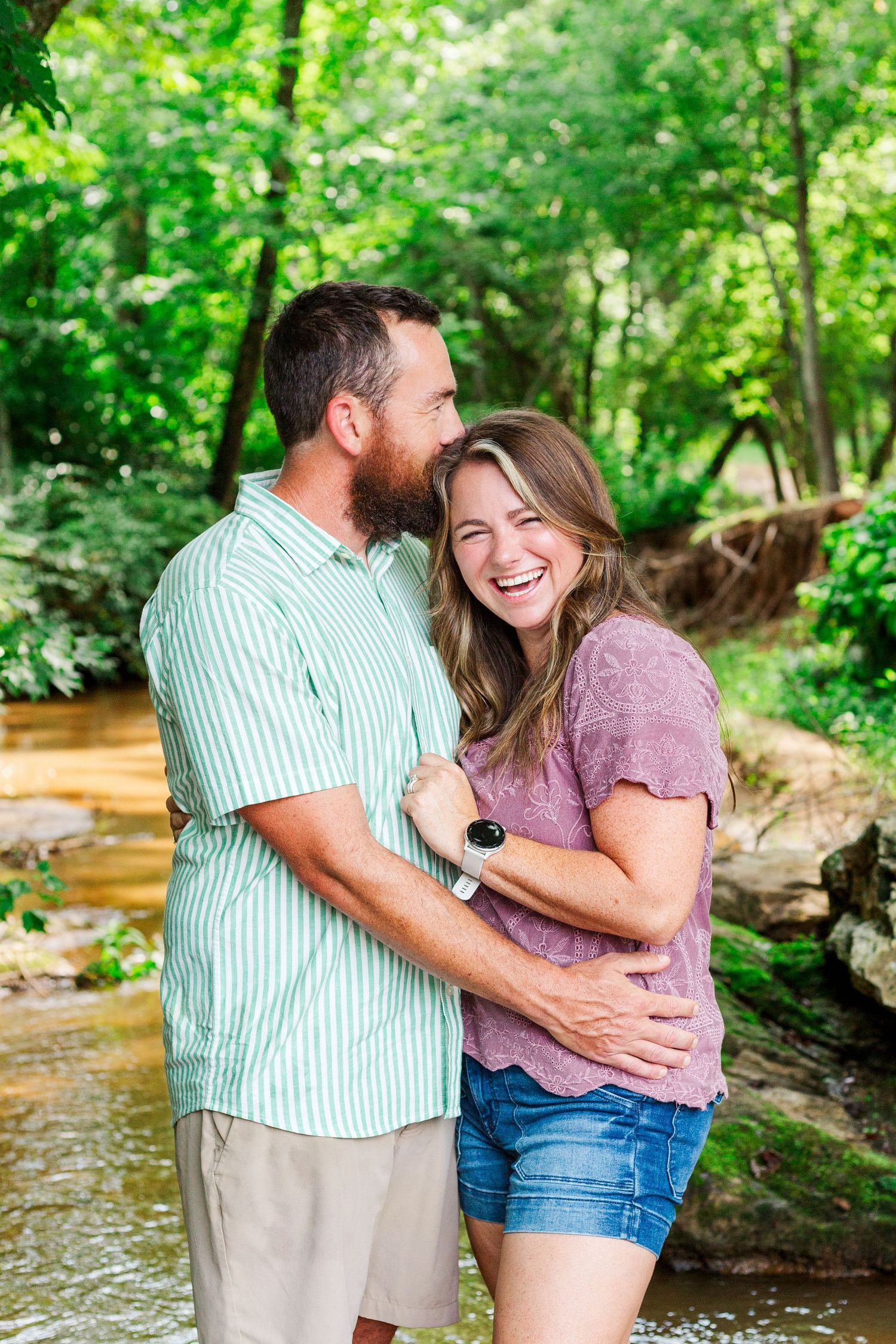 A couple shares tender moments and laughter by a creek surrounded by green summer foliage.
