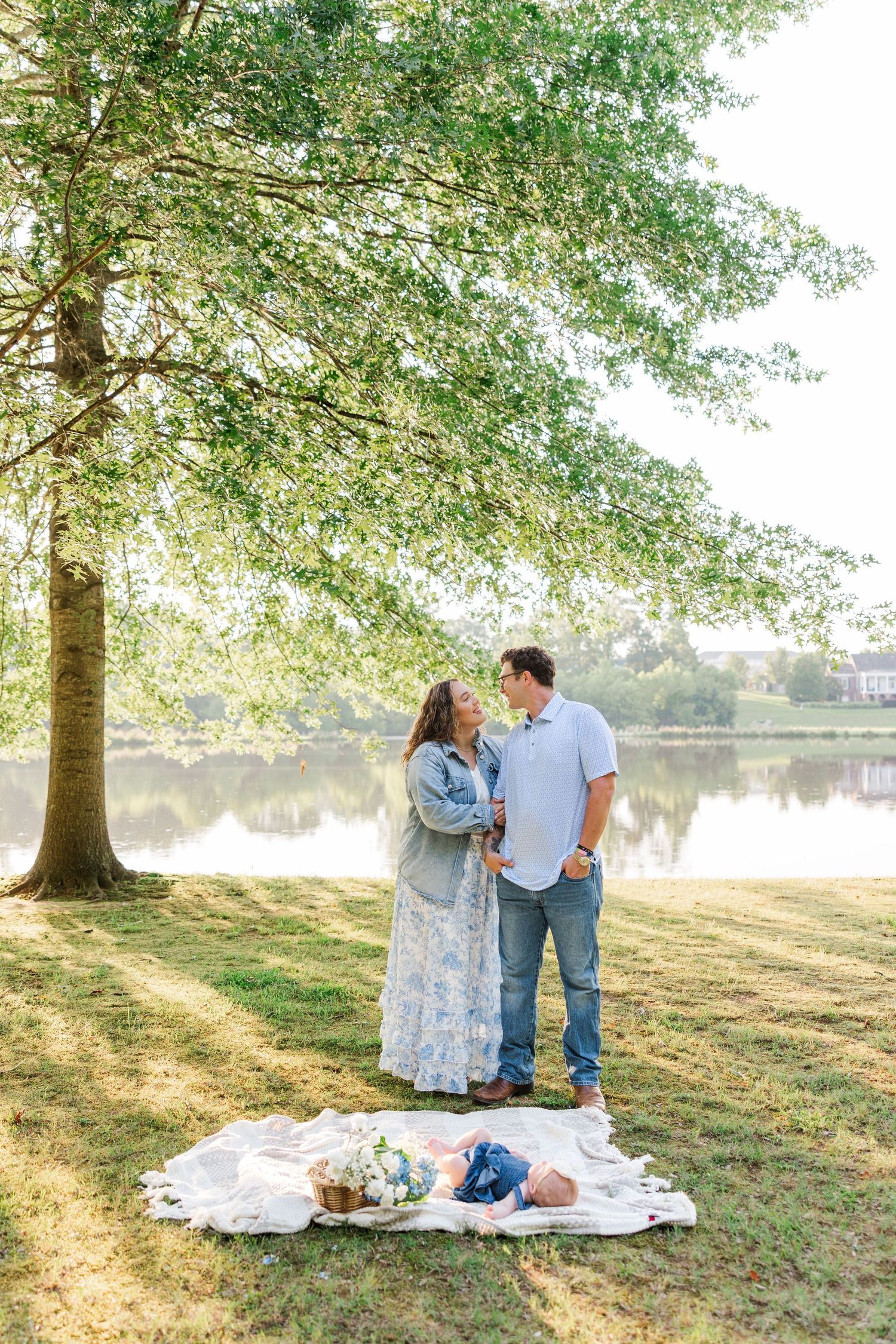 Couple sharing a tender moment by a serene lake under a large tree, with a baby resting on a blanket nearby.