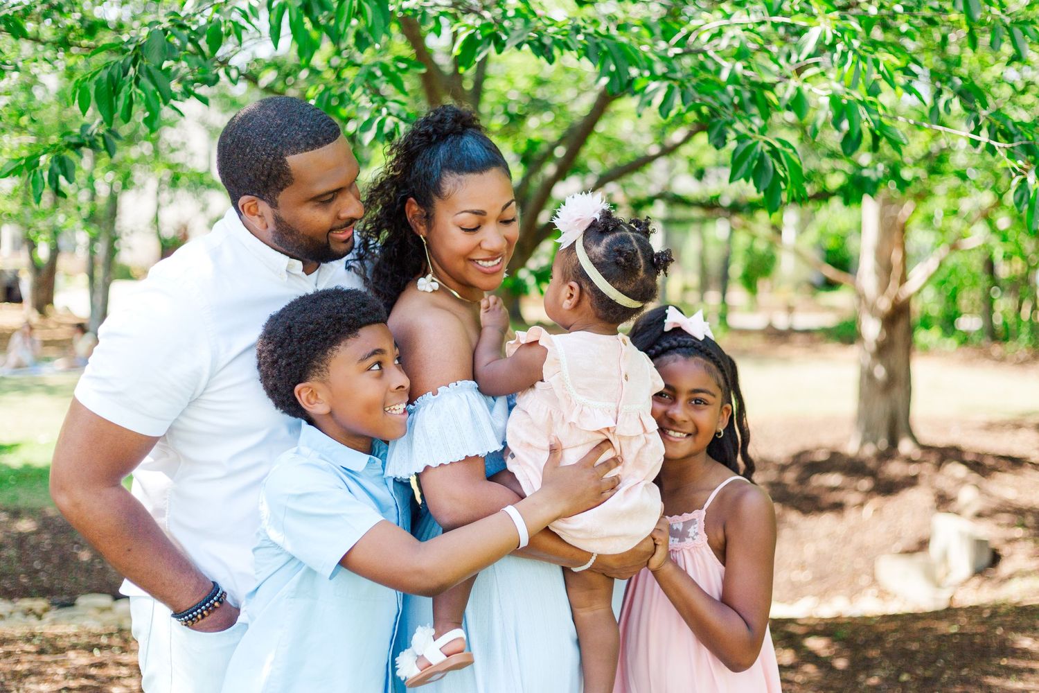 Happy family with two kids and a baby smiling together outdoors under green trees in a park.