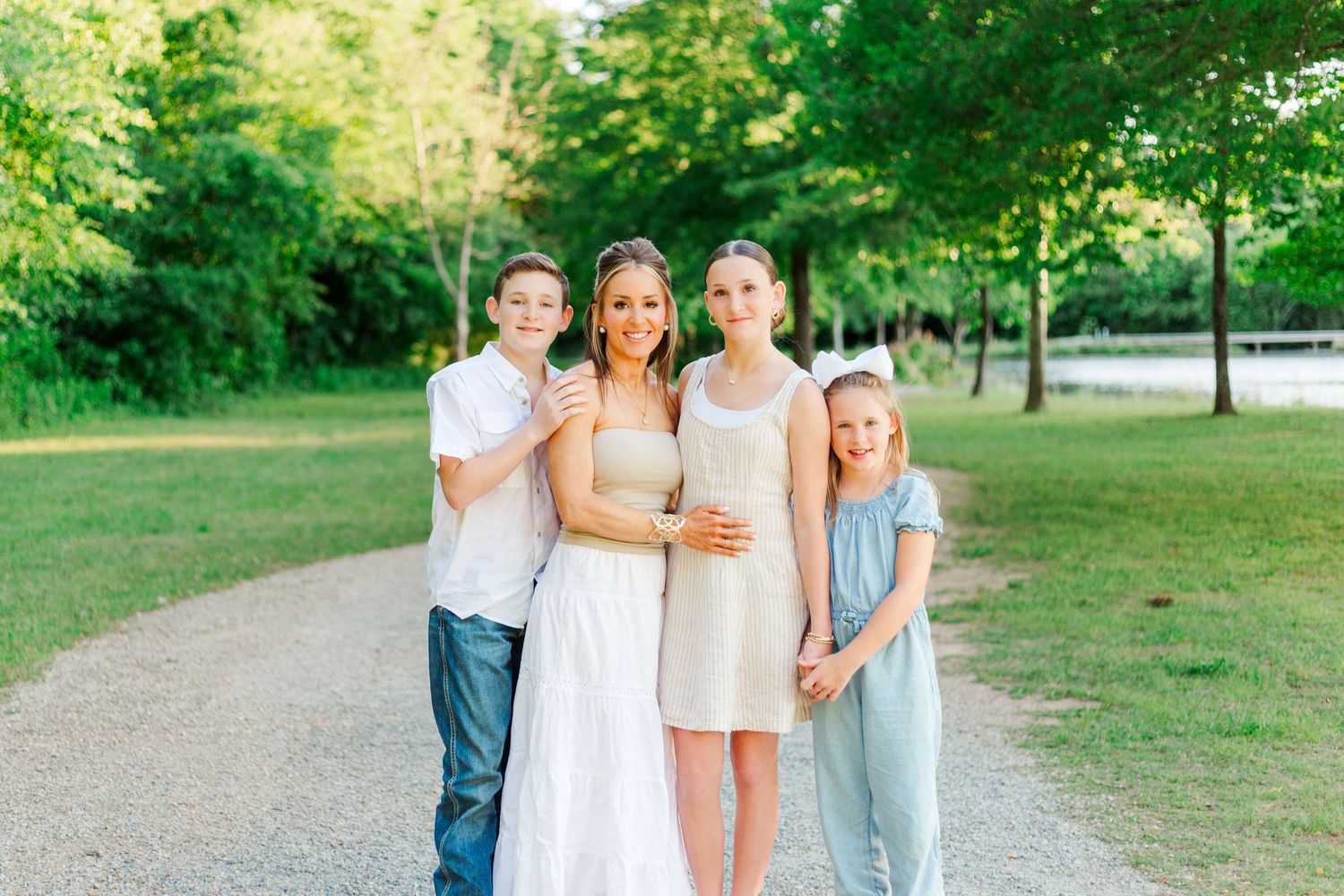 Family portrait of a woman in white dress with three children smiling on a park path surrounded by green trees.