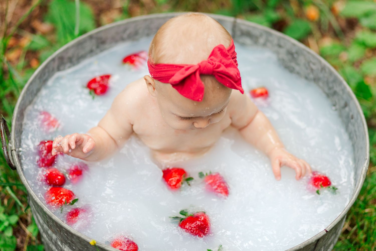 A baby wearing a red bow sits in a metal tub filled with bubbles and scattered red flowers.
