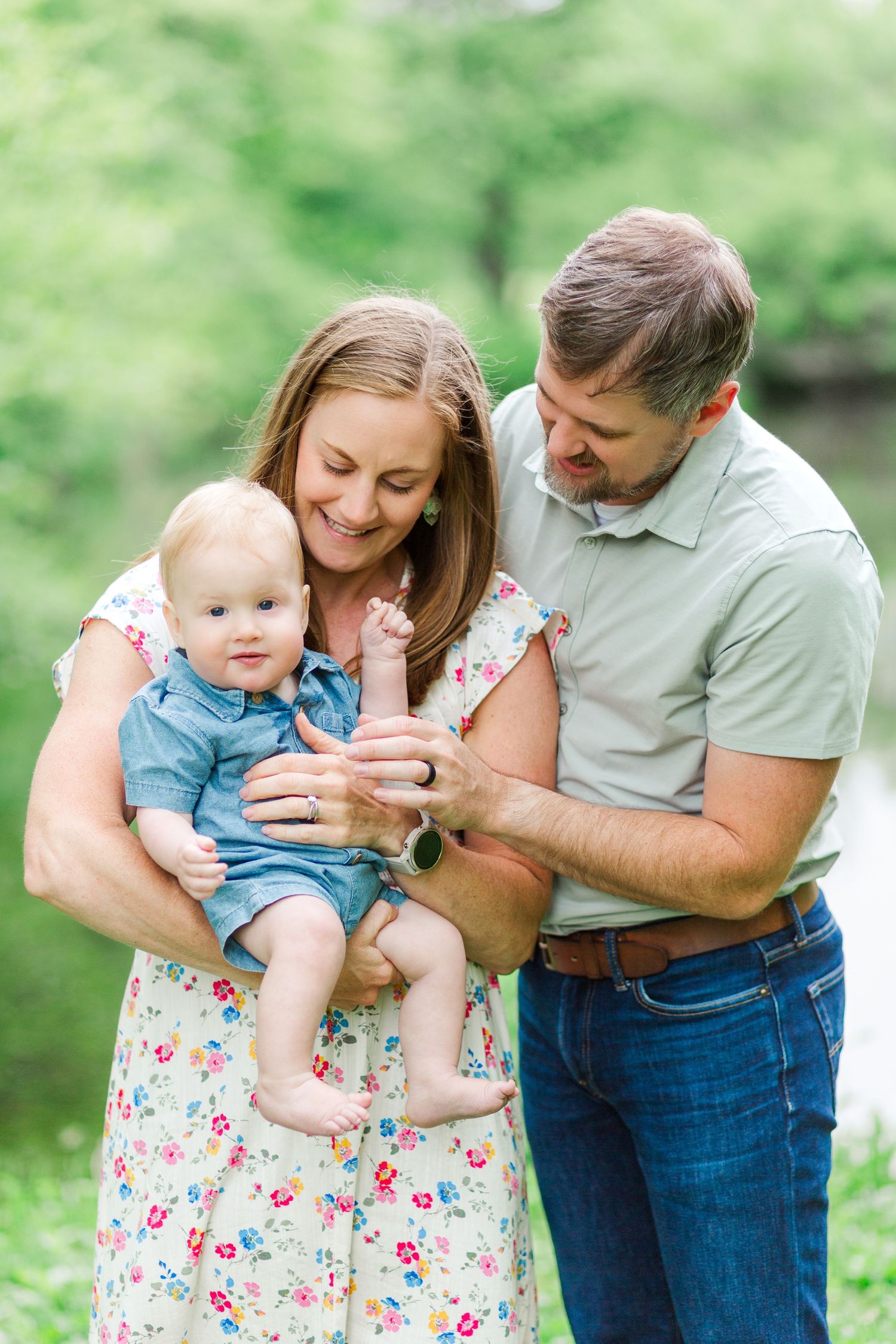 A smiling couple holds their baby boy outdoors in a lush green park during a family portrait session.