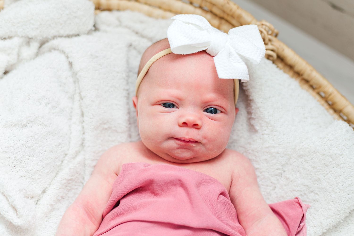 A baby wearing a white bow headband lies on a white blanket in a woven basket.