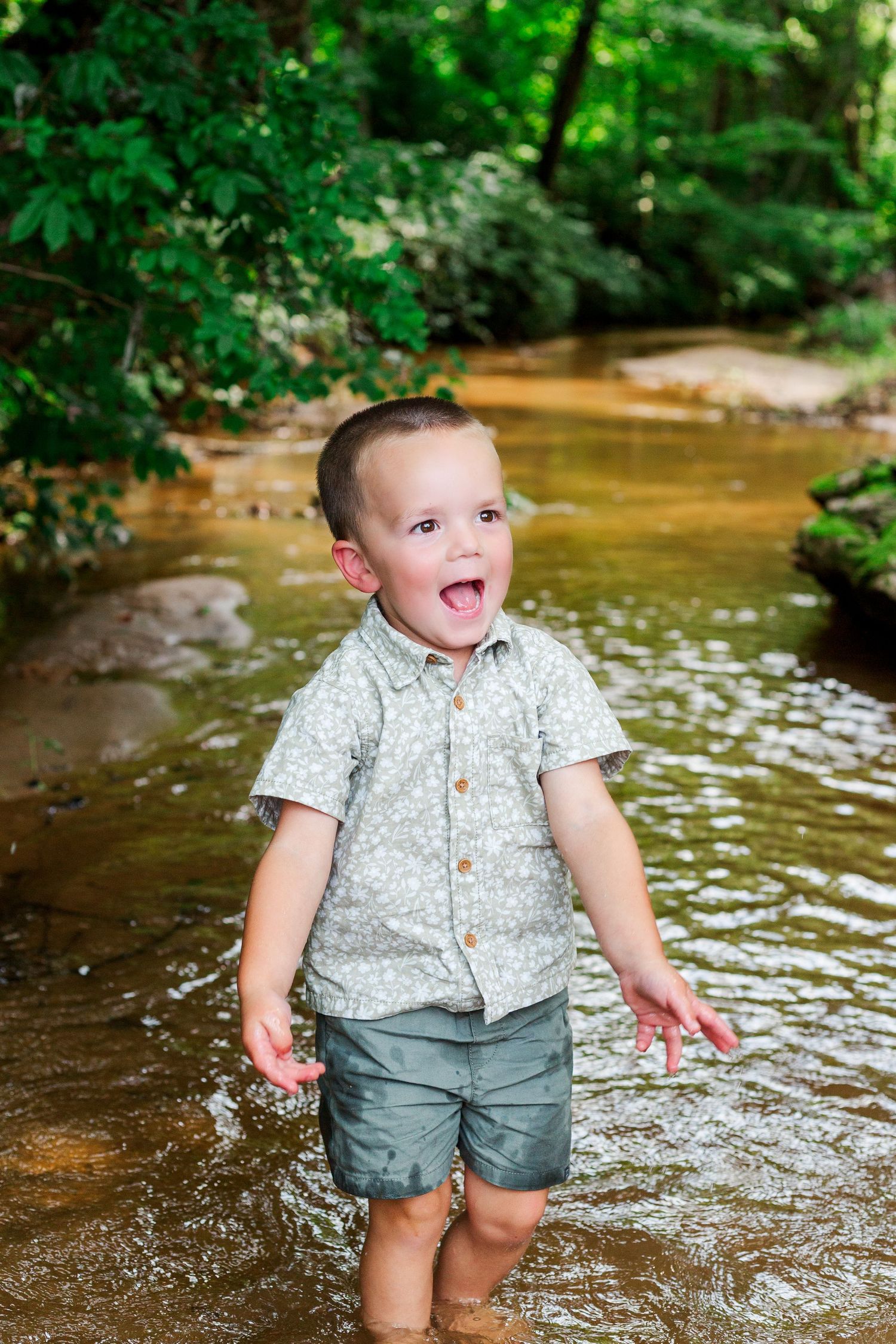 Small child splashes and plays in a creek wearing a patterned shirt and gray shorts.