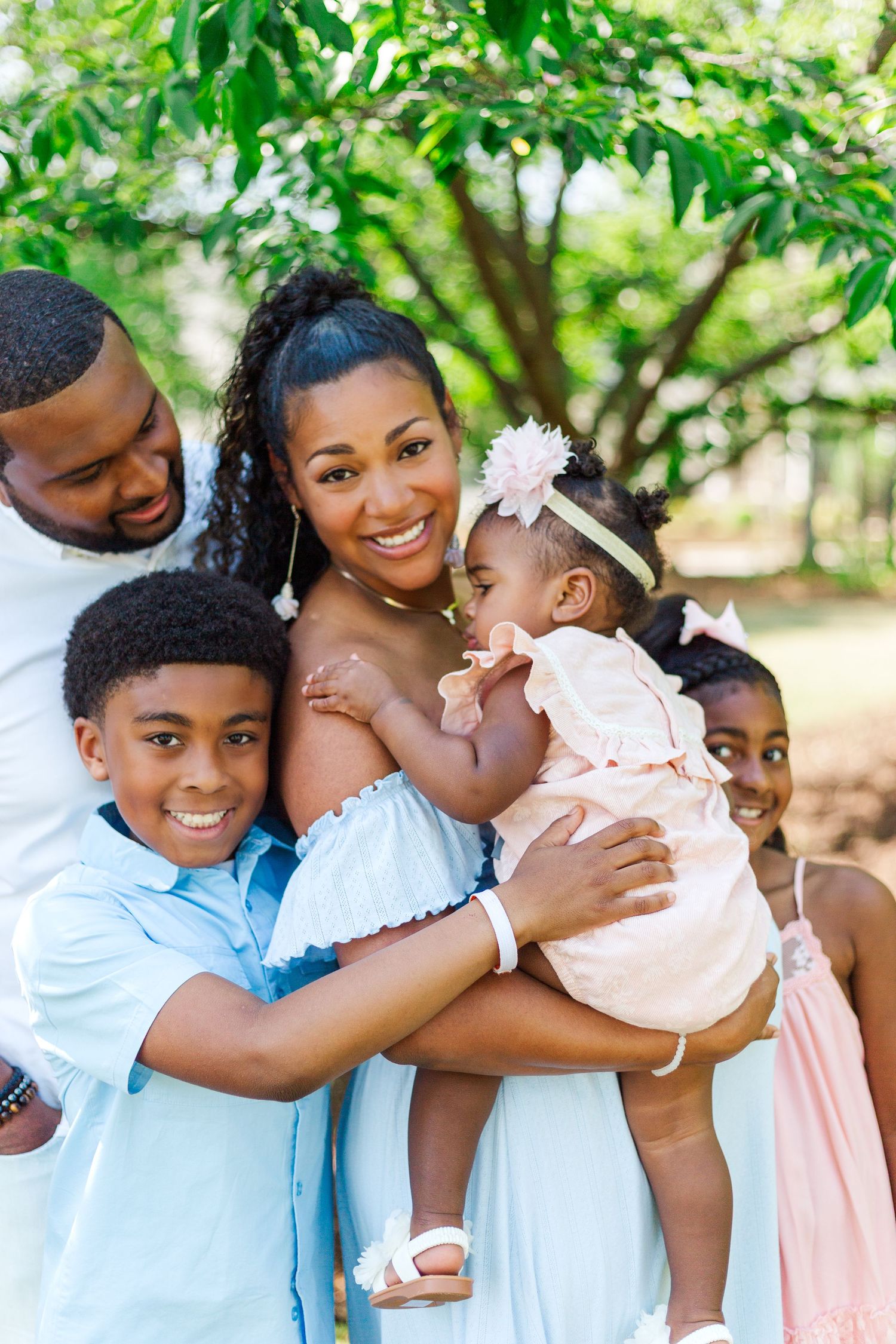 Happy Black family posing outdoors under green trees, parents with three children in coordinated pastel outfits.