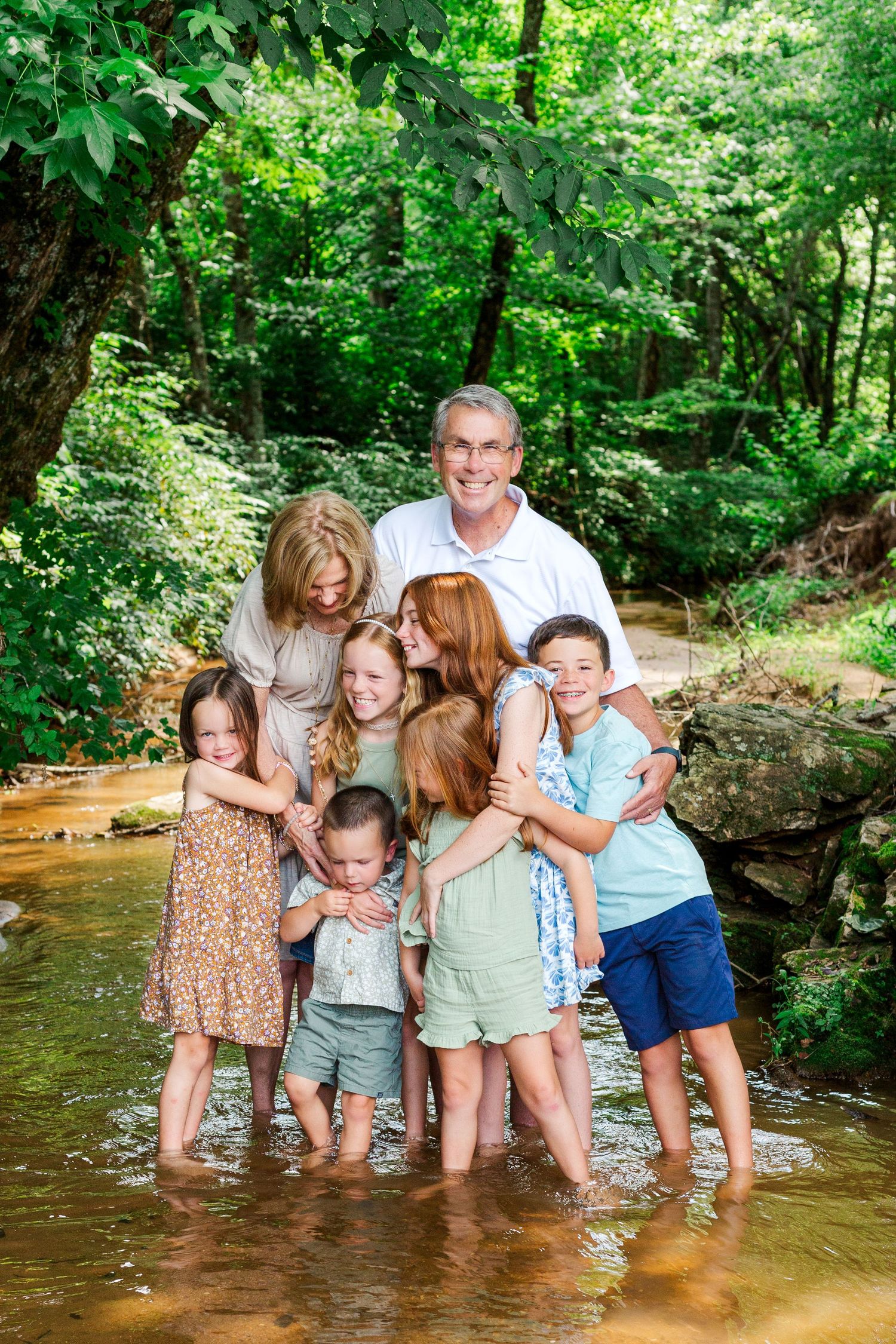A group stands together in a shallow creek surrounded by lush green forest on a bright summer day.