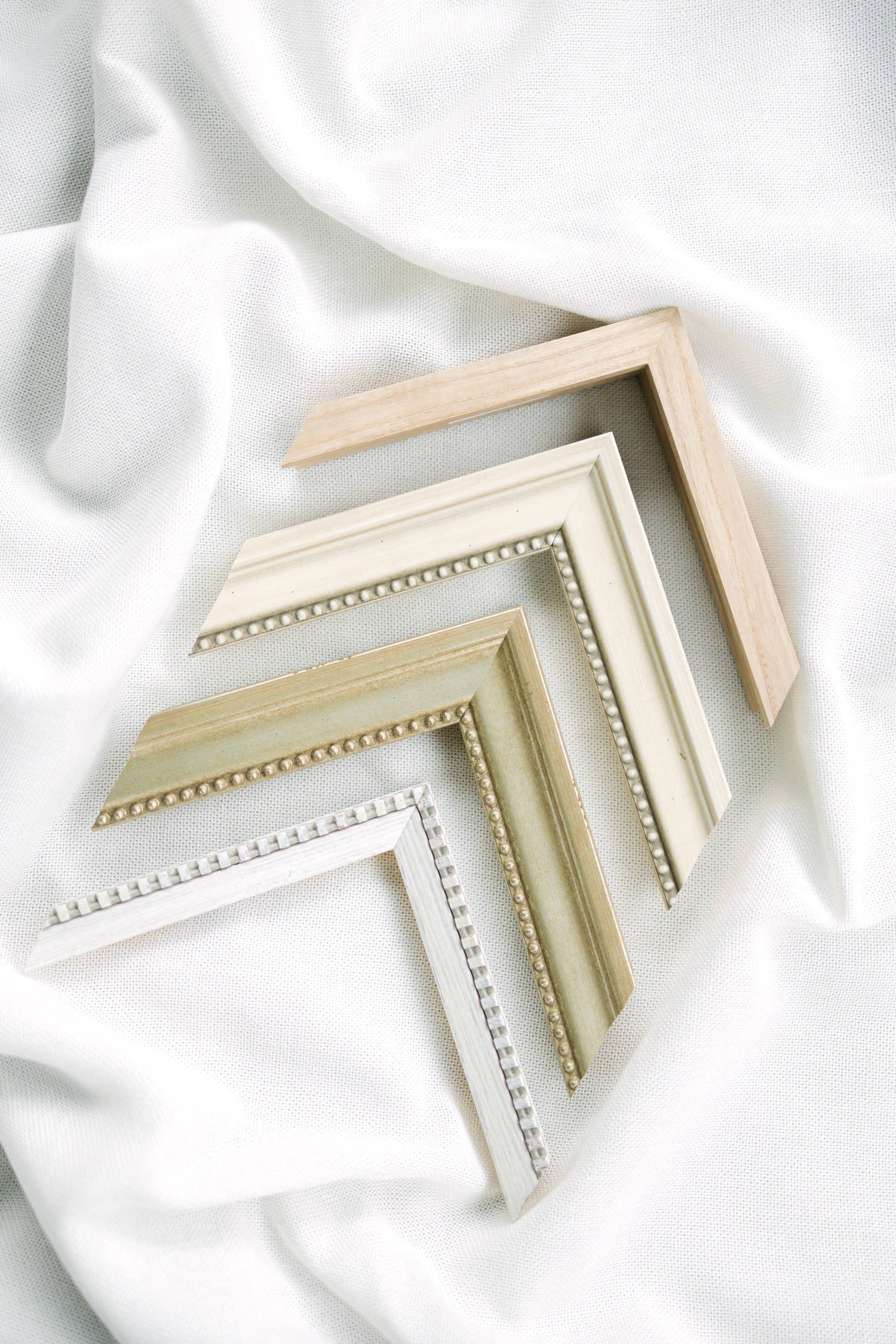 Four decorative picture frame corners in neutral tones arranged on white silk fabric.