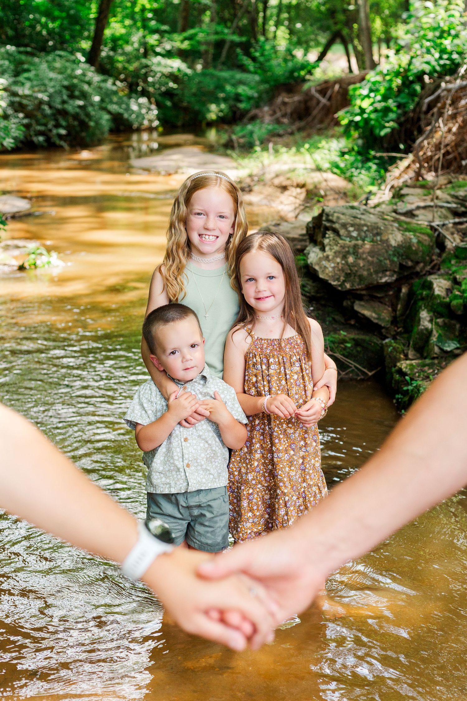 Three children standing in a creek with holding hands visible in foreground on a sunny summer day.
