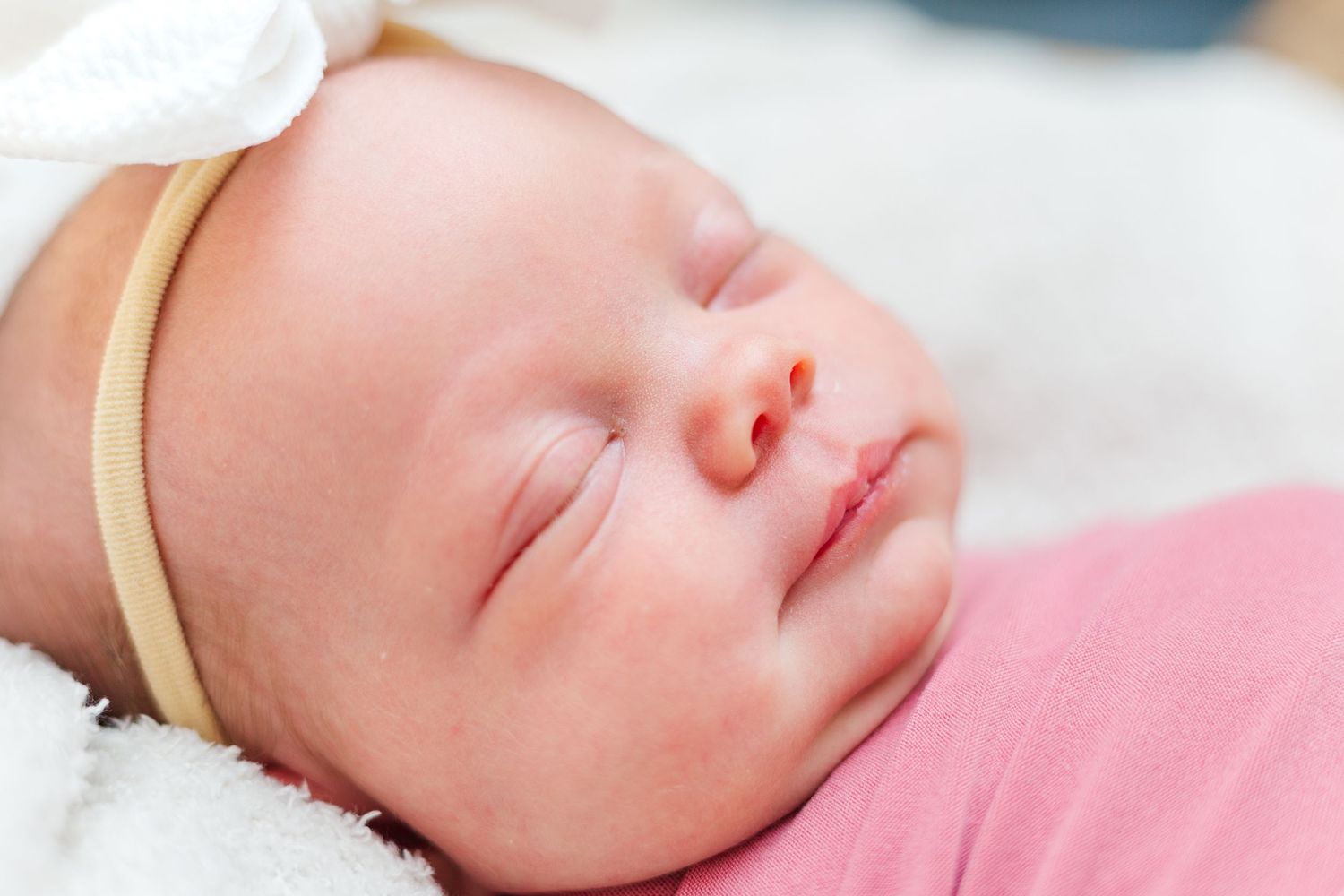 A sleeping newborn baby in a pink outfit rests peacefully on a white blanket with a thin headband.