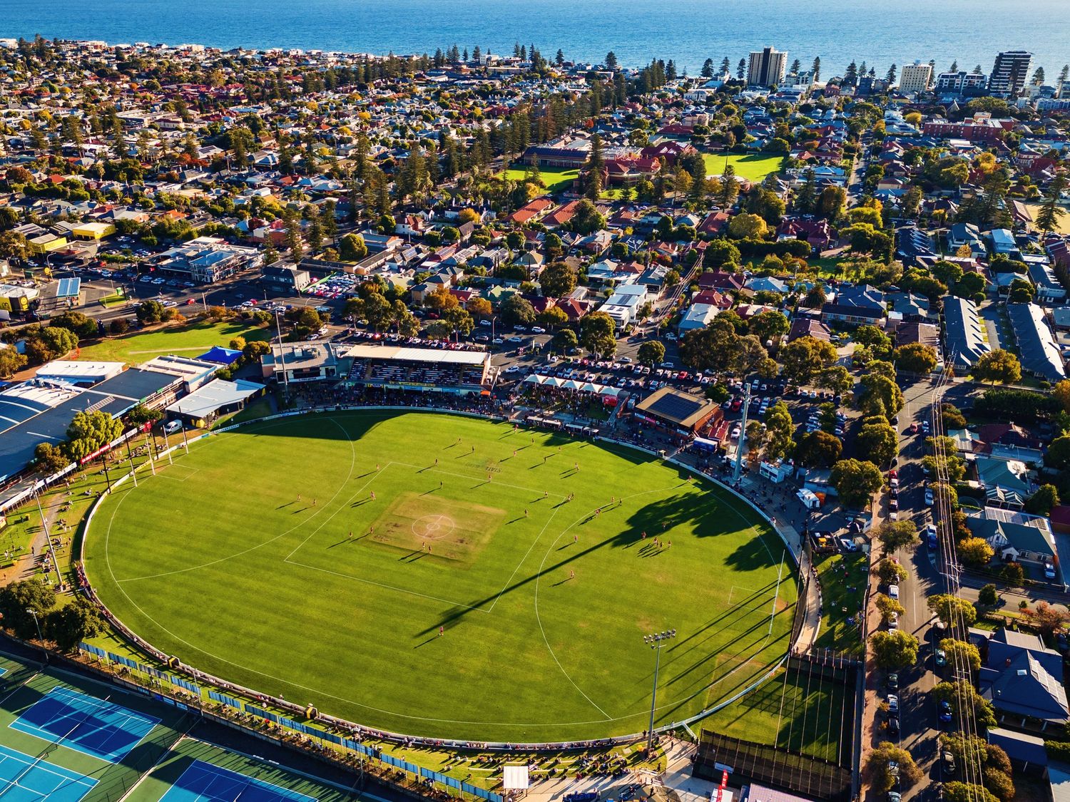 Aerial view of oval cricket ground surrounded by suburban houses with city skyline in background.