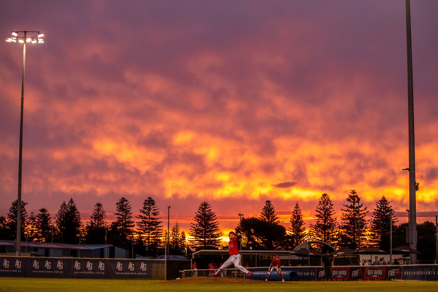 Vibrant orange and purple sunset sky over a sports stadium with dark silhouetted buildings.