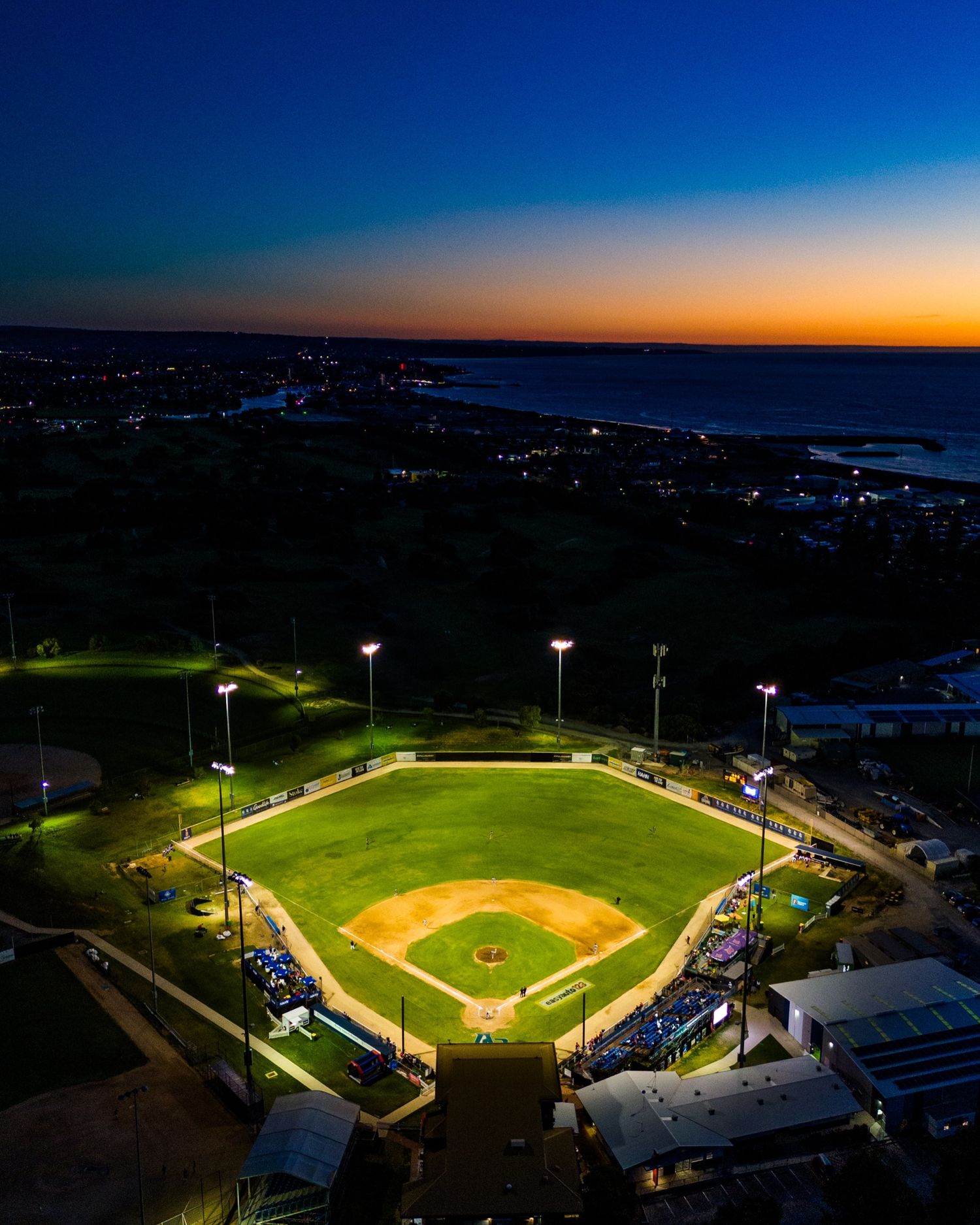 Aerial view of an illuminated baseball stadium at dusk with lights glowing against dark sky.