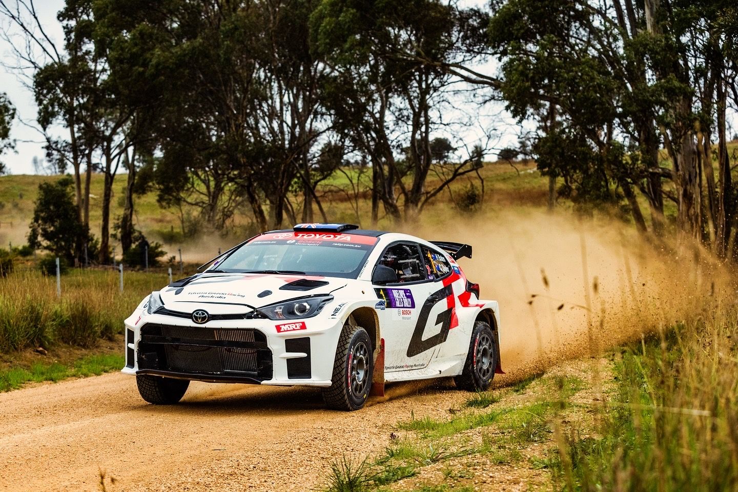 White rally car kicks up dust while racing on dirt road through Australian bushland.