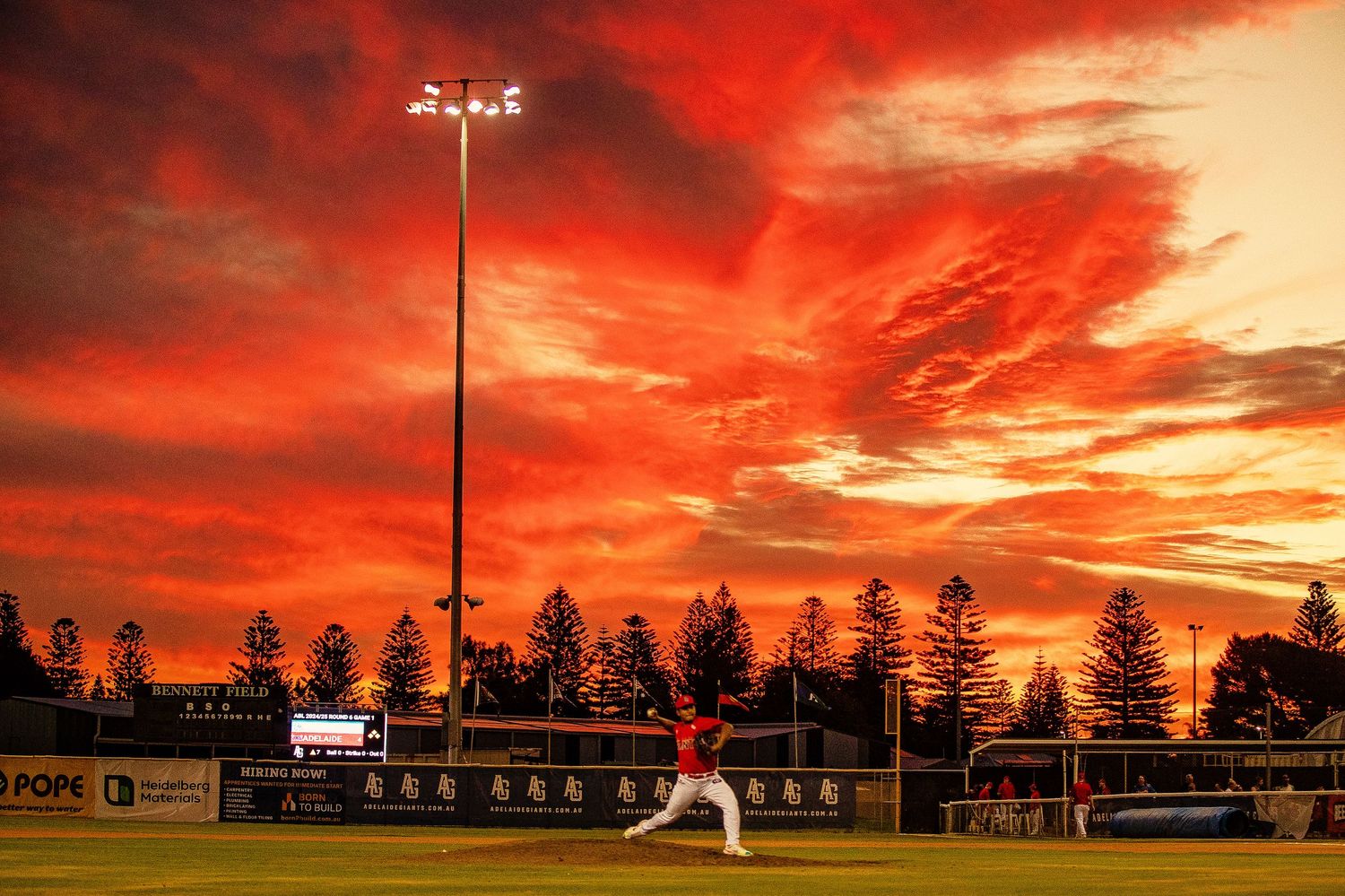 Dramatic red and orange sunset sky over a sports field with silhouetted trees in the background.