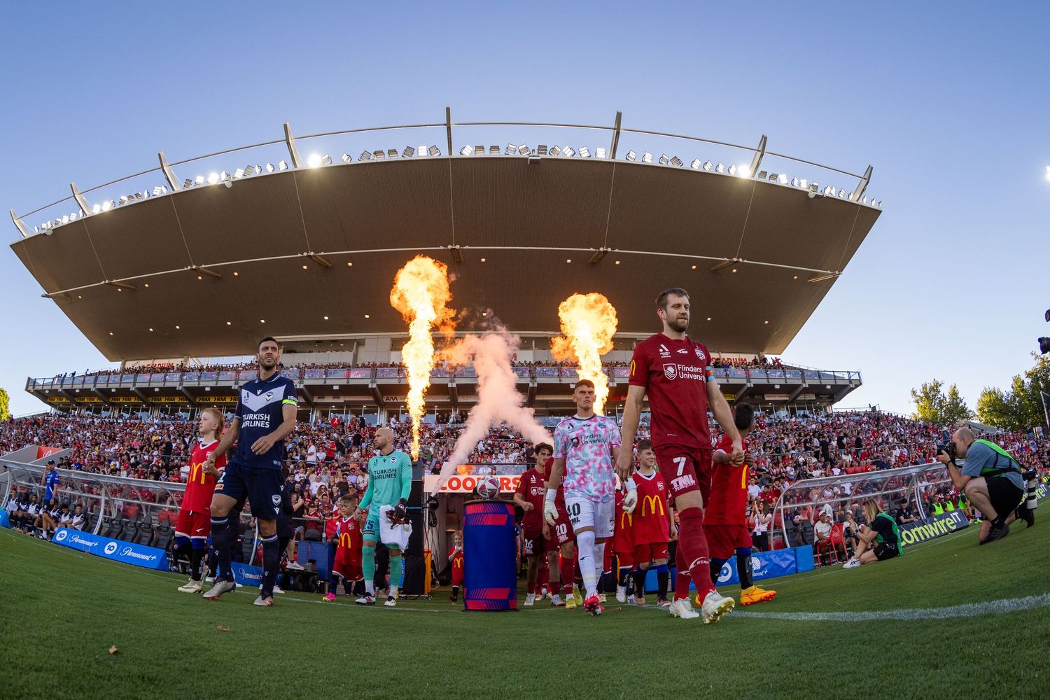 Panoramic view of a sports stadium with crowd and flaming pyrotechnics at sunset.