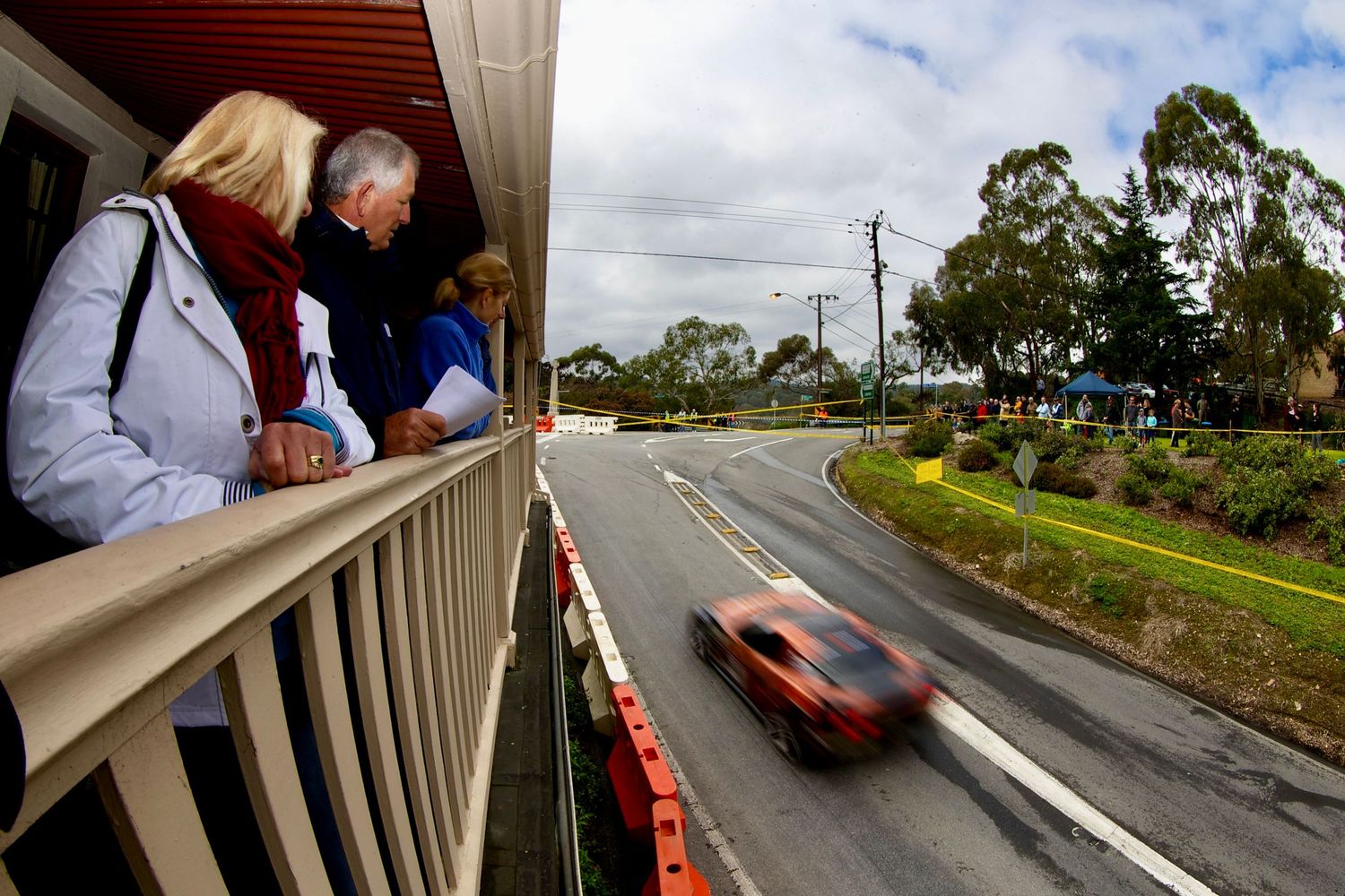 Spectators watch from balcony as blurred car races past on suburban street.