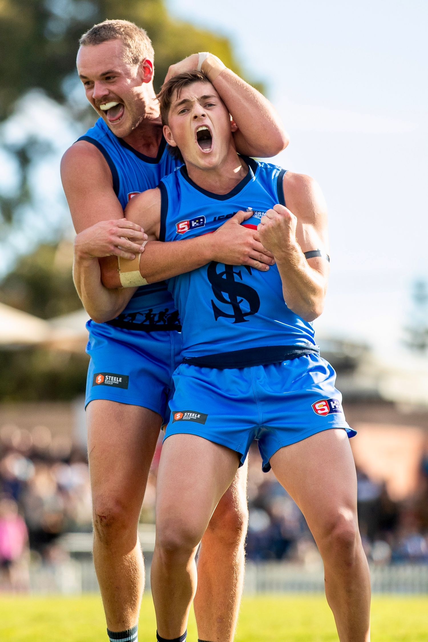 Athletes in blue uniforms celebrate victory with joyful embraces on sunny field.