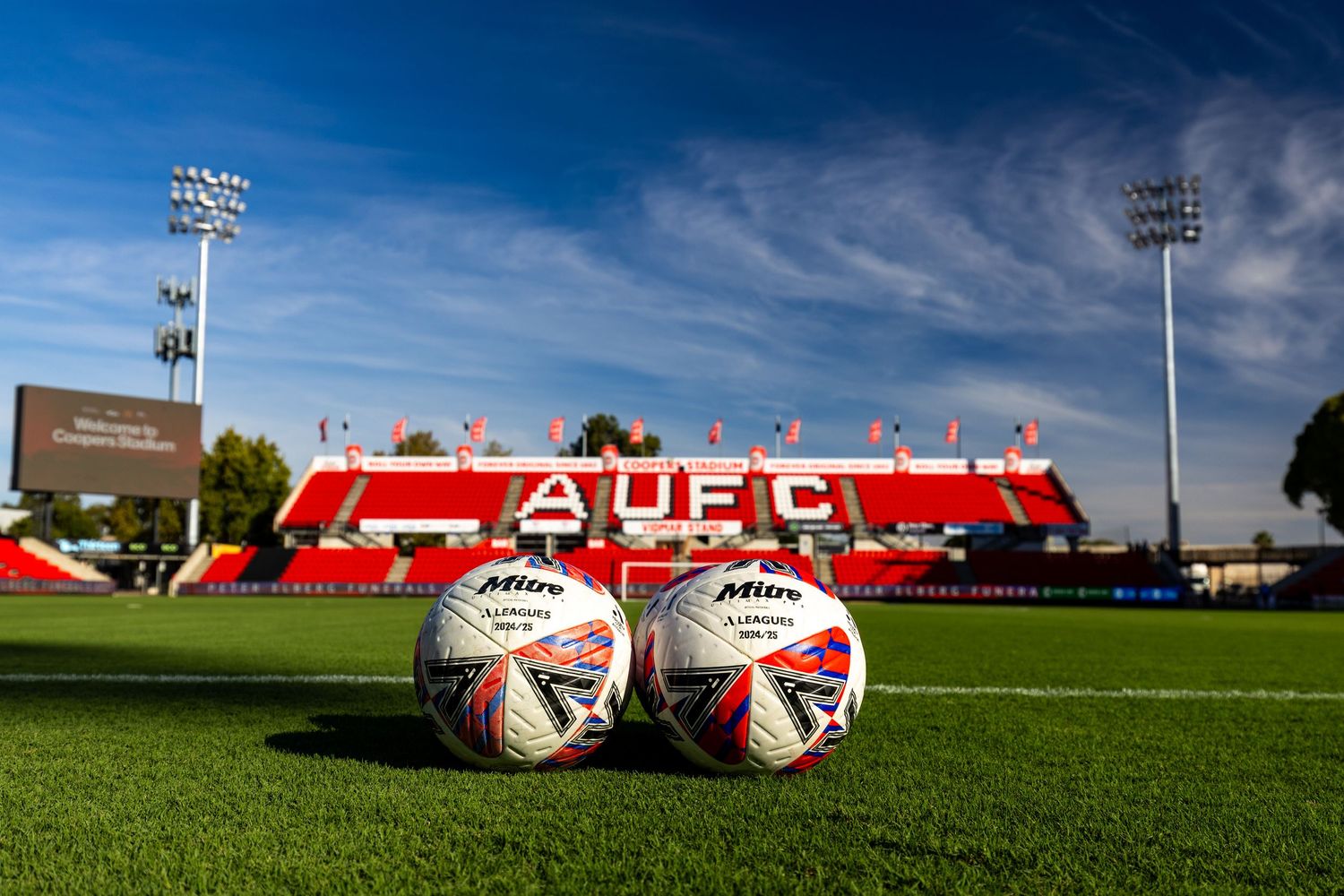 Two soccer balls rest on green grass with red stadium seats in background under blue sky.