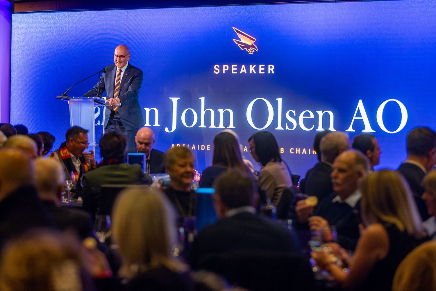 Conference stage with blue lighting and speaker text displayed on large screen.
