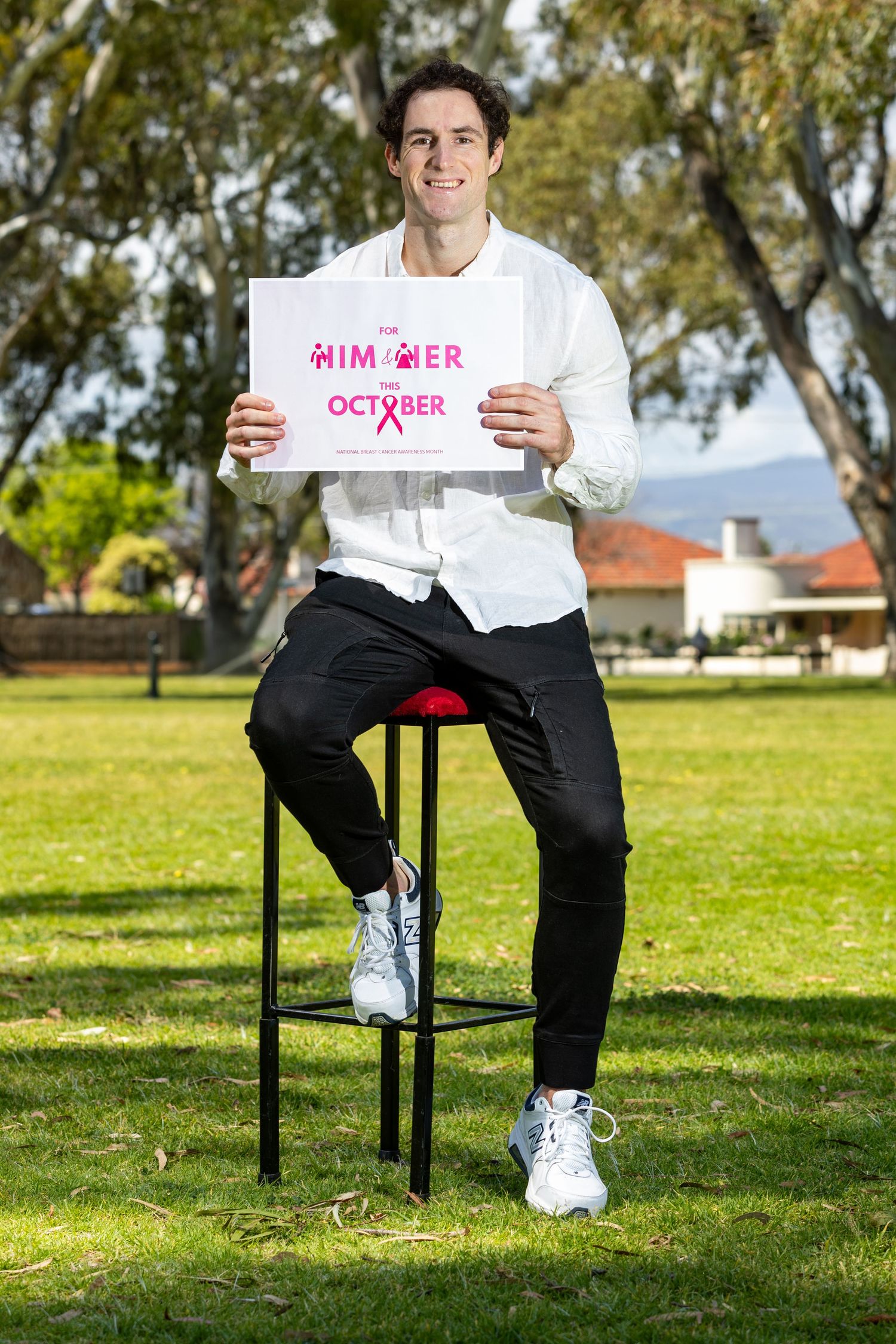 Person holds pink breast cancer awareness sign while sitting on stool outdoors.