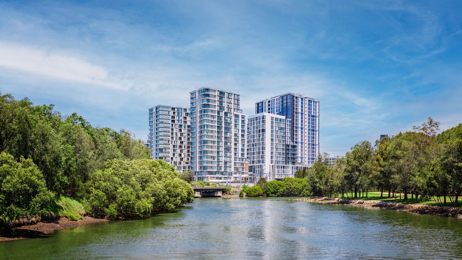 Panoramic view of modern high-rise buildings along a waterfront with lush green trees and blue sky.