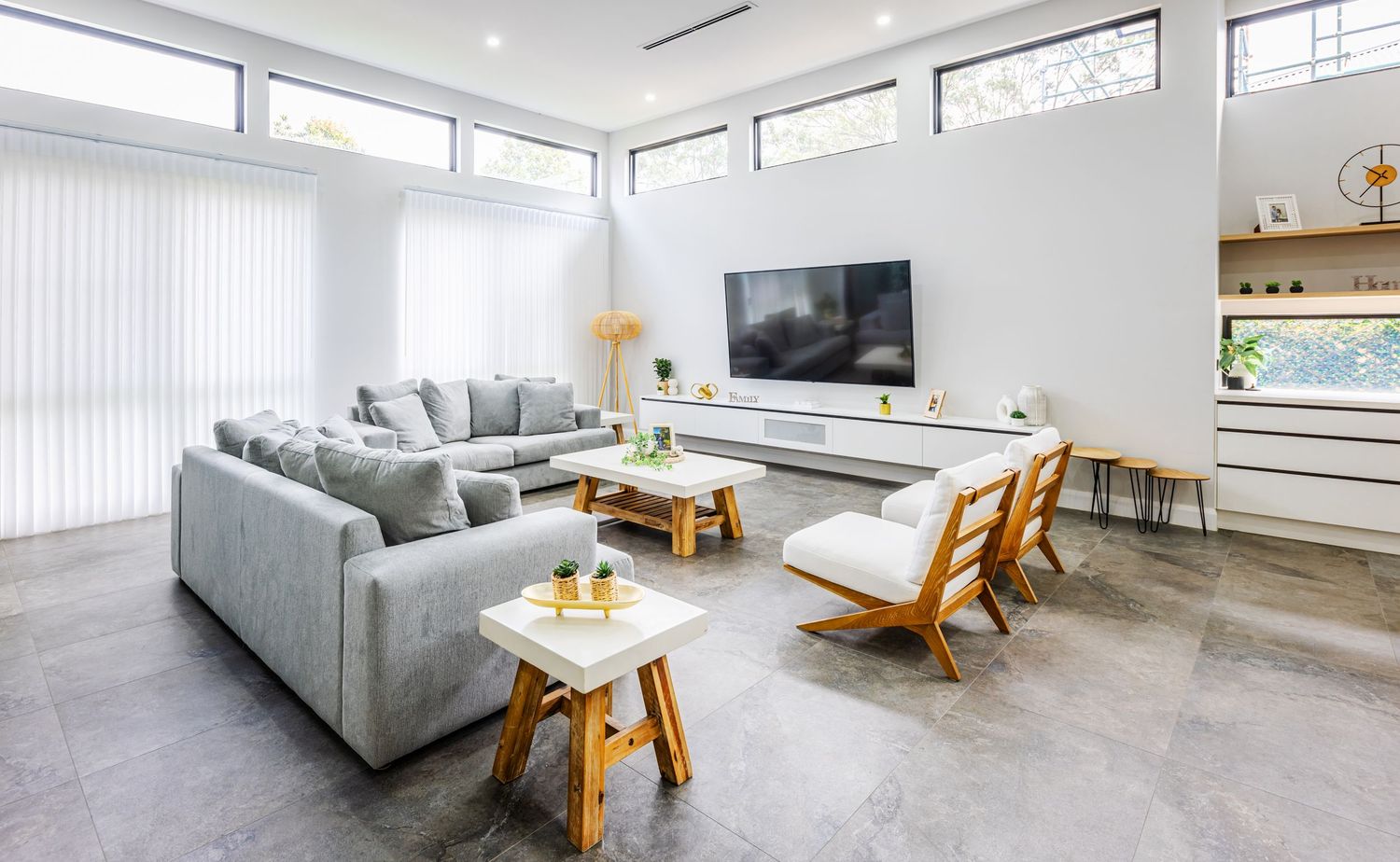 Modern living room with gray sectional sofa, wooden accent chairs, and wall-mounted TV in bright white space.