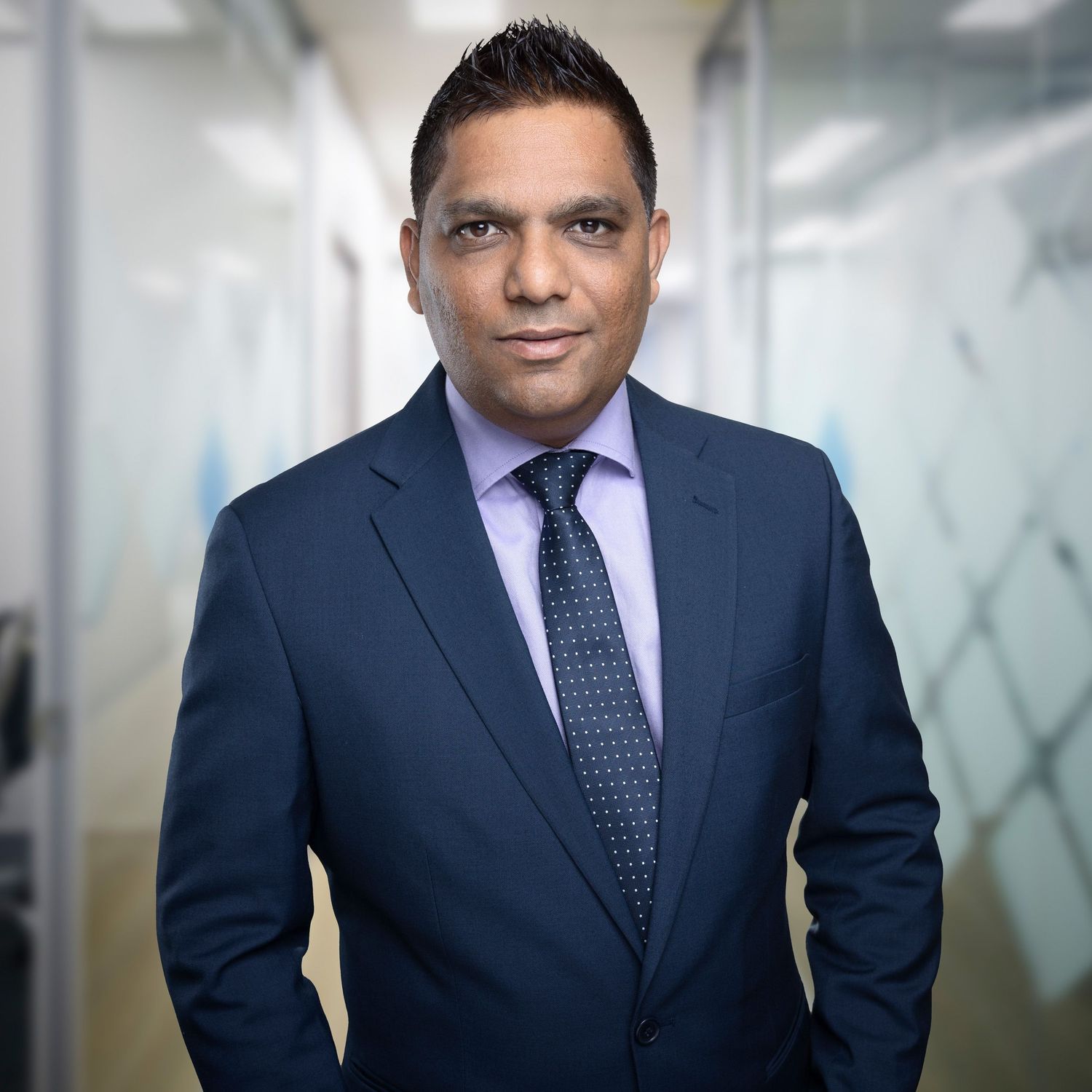 Professional business portrait of person in navy suit and purple tie against blurred office background.